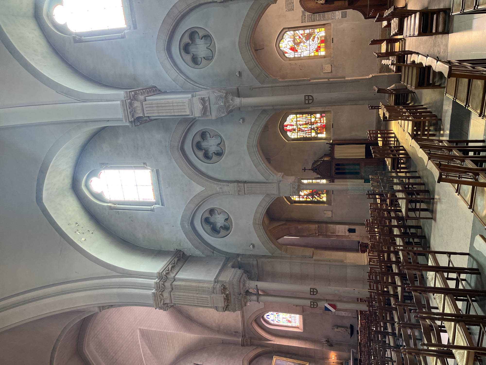 The image depicts the interior of the Église Saint-Lubin in Rambouillet, France. The church features a high vaulted ceiling with Gothic arches. Sunlight streams through stained-glass windows, casting colorful patterns on the stone walls and floors. Rows of wooden chairs, typical of church seating, are arranged in the nave. Architectural elements include stone columns, decorative carvings, and circular quatrefoil windows above arched openings. A confessional booth is visible on the left, and a French flag hangs on a column in the distance. The overall impression is one of solemnity and architectural grandeur.