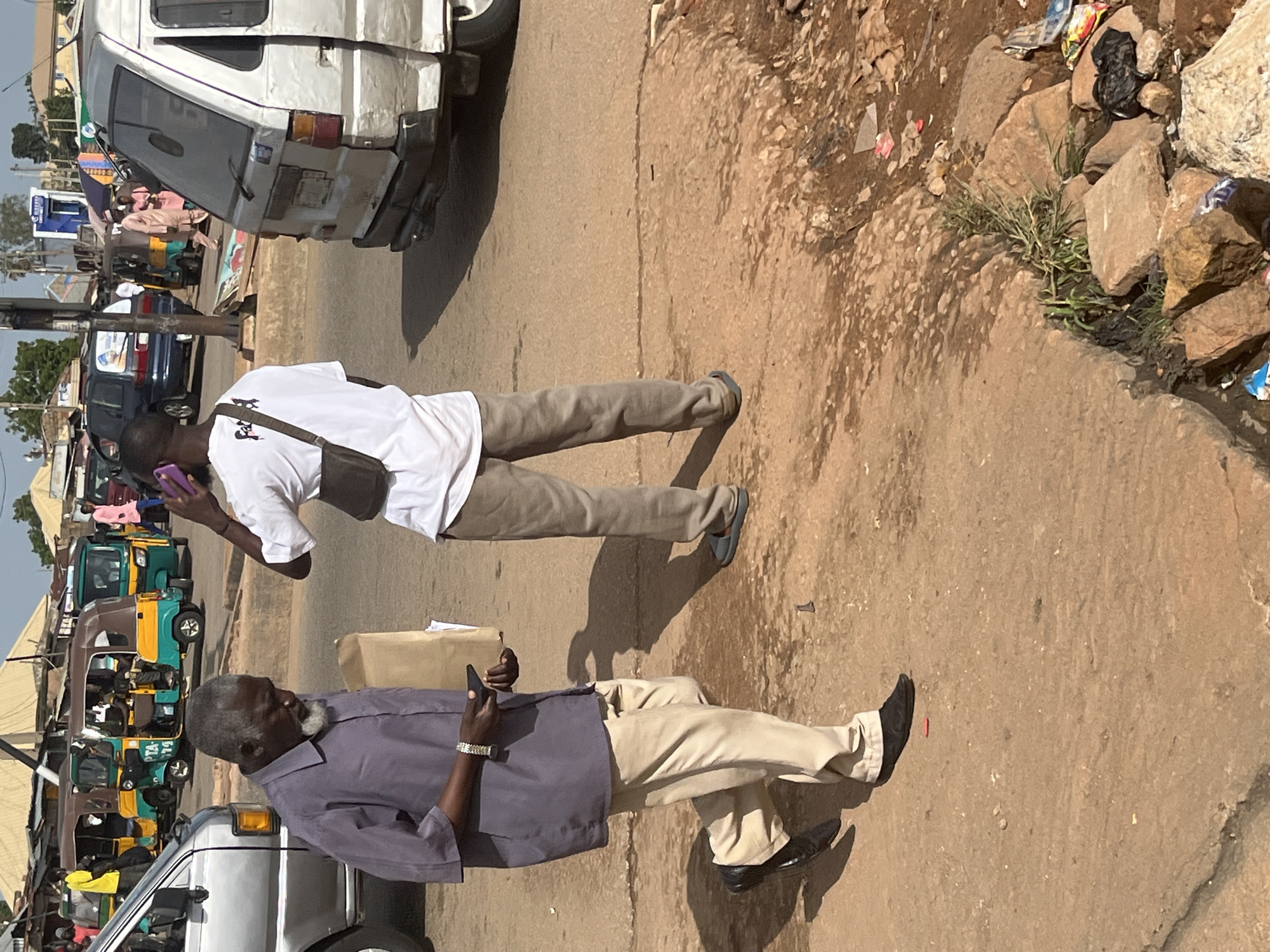 The image depicts a street scene in Gura Top, Nigeria. In the foreground, an older Black man with a grey beard and mustache walks from left to right, wearing a dark purple collared shirt, light beige trousers, and black dress shoes. He holds a light brown paper bag in his left hand and a black mobile phone in his right hand, which he appears to be looking at. To his right, another Black man stands, facing away from the camera, wearing a white t-shirt with a dark graphic and beige trousers. He is holding a purple mobile phone to his ear. In the background, various vehicles are visible, including a white van on the right, several colorful auto-rickshaws (Keke Napeps), and a dark blue SUV. People can be seen walking and standing around the vehicles and market stalls, suggesting a bustling urban environment. The ground is paved with dirt and some rubble, with patches of sparse vegetation. The overall impression is of a typical day in a busy Nigerian town.