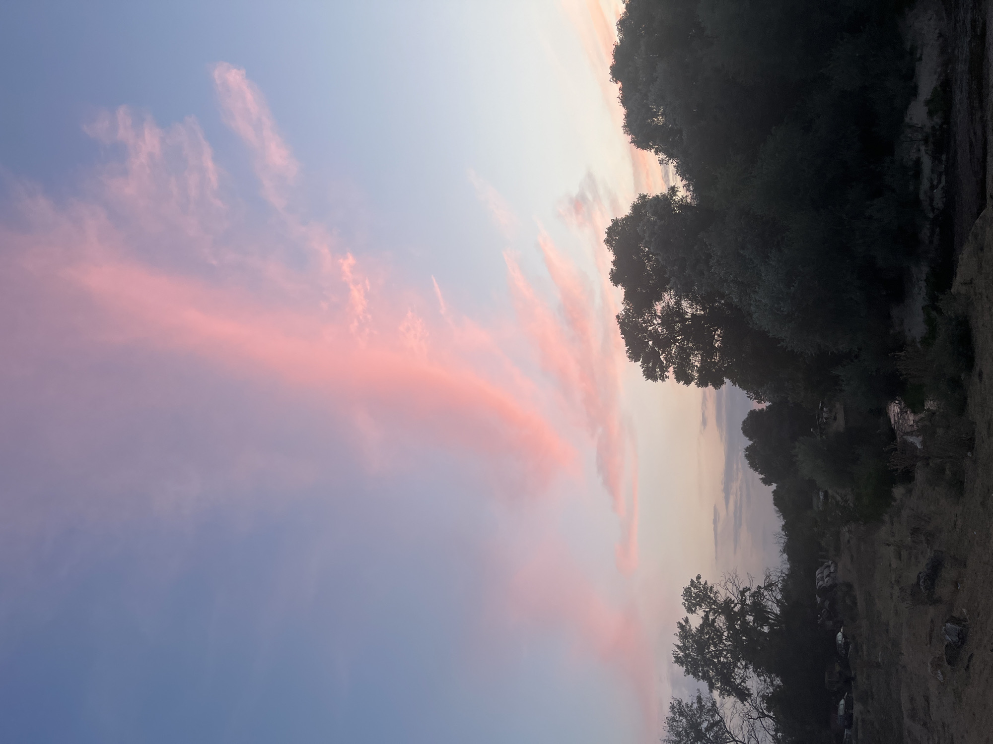 This image depicts a landscape scene at dusk, featuring a vibrant sky with pink and blue hues, likely from a sunset. The sky is streaked with wispy clouds that are illuminated by the setting sun, creating a striking visual effect.
In the foreground, a natural landscape is visible, characterized by a mix of trees and scrubland. The trees are silhouetted against the sky, suggesting they are dense and dark. The ground appears dry and uneven, with patches of dirt and possibly some rocks. There are also hints of what might be a body of water or a dry riverbed in the lower right portion of the image, reflecting the subtle colors of the sky.
The presence of scattered objects in the mid-ground, particularly in the left side of the image, suggests human activity and a potential junkyard or abandoned area. These objects include what appear to be several vehicles, some of which are partially obscured by vegetation. There are also stacks of round items, possibly tires or large containers. This element introduces a contrast between the natural beauty of the sky and the more industrial or neglected aspect of the land.
The location is specified as Plovdiv, Bulgaria. While the image itself doesn't provide specific landmarks to pinpoint an exact spot within the city, the overall depiction is consistent with a peri-urban or rural fringe area where natural landscapes meet signs of human development or decay. The scene captures a moment of natural beauty juxtaposed with evidence of human presence.