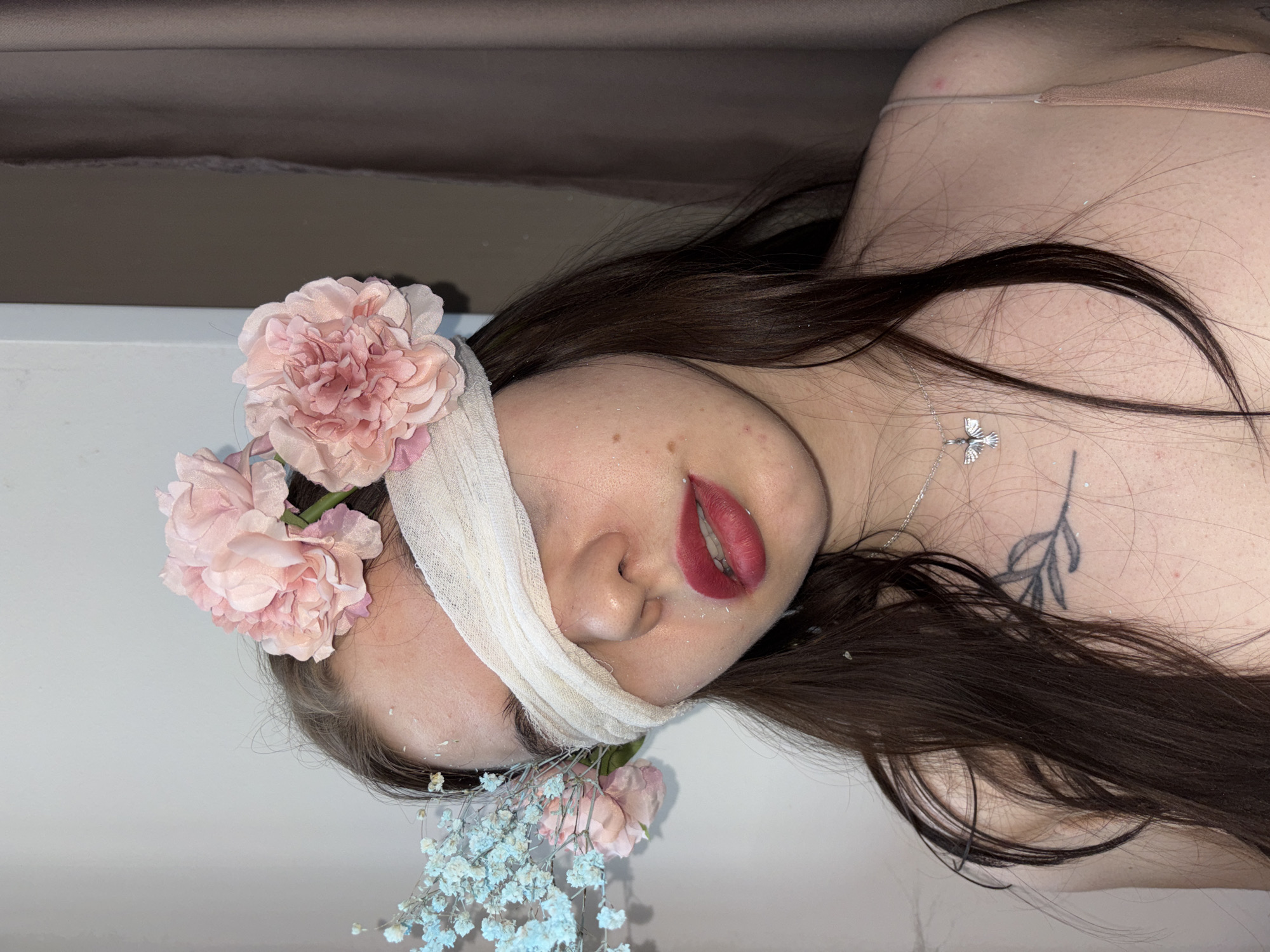 This media file features a close-up, vertically oriented shot of a person, likely female, lying down indoors. The individual's eyes are covered with a light-colored, textured cloth bandage or wrap that extends across the bridge of the nose. Her lips are painted with dark red lipstick and are slightly parted.
Long, dark brown hair is spread around her head and shoulders. Artificial light pink flowers, resembling carnations or peonies, are arranged near her left temple. A cluster of artificial light blue baby's breath flowers is visible near her chin and neck. Fine white powder or glitter is scattered on her skin and hair.
A silver necklace with a small, detailed dove pendant rests on her upper chest. A minimalist tattoo depicting a leafy branch is visible on her left shoulder area. A light-colored bra strap is also visible on her right shoulder. The background consists of a plain, light surface in the foreground and a darker, blurred area behind the subject. The image suggests a posed, artistic portrait. No visible text is present. The context provided for this image is Perm, Russia.