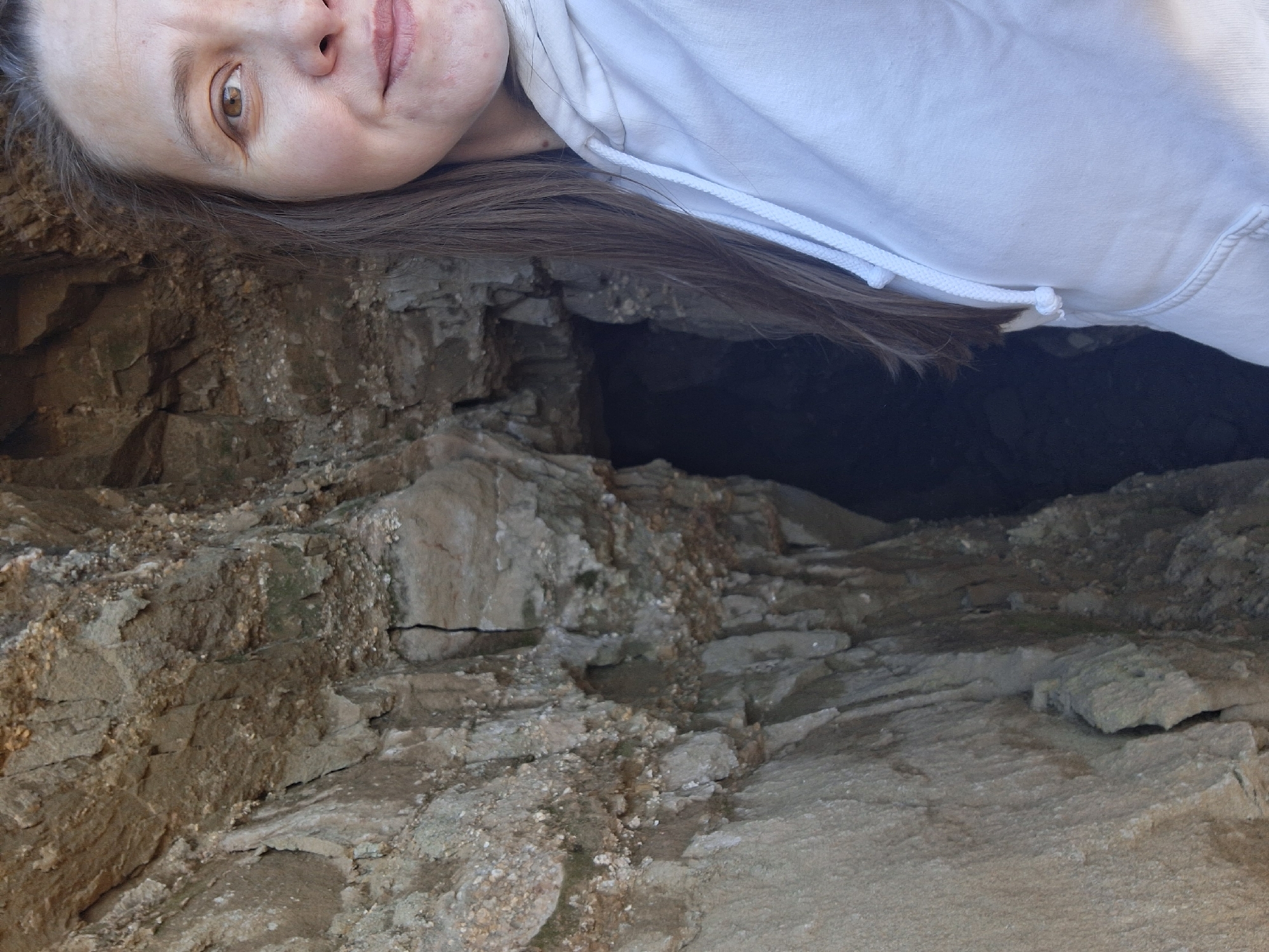 A young woman lies near a cave entrance in what appears to be a rocky, possibly eroded, area. The cave opening is dark, hinting at depth. The woman's head is close to the cave entrance, suggesting she may be looking inside. She is wearing a white hoodie. The location is likely Chaikovsky, Russia, if that information is reliable from the prompt. The overall impression is of exploration or curiosity in a natural setting. The image is presented in an unusual, rotated perspective.