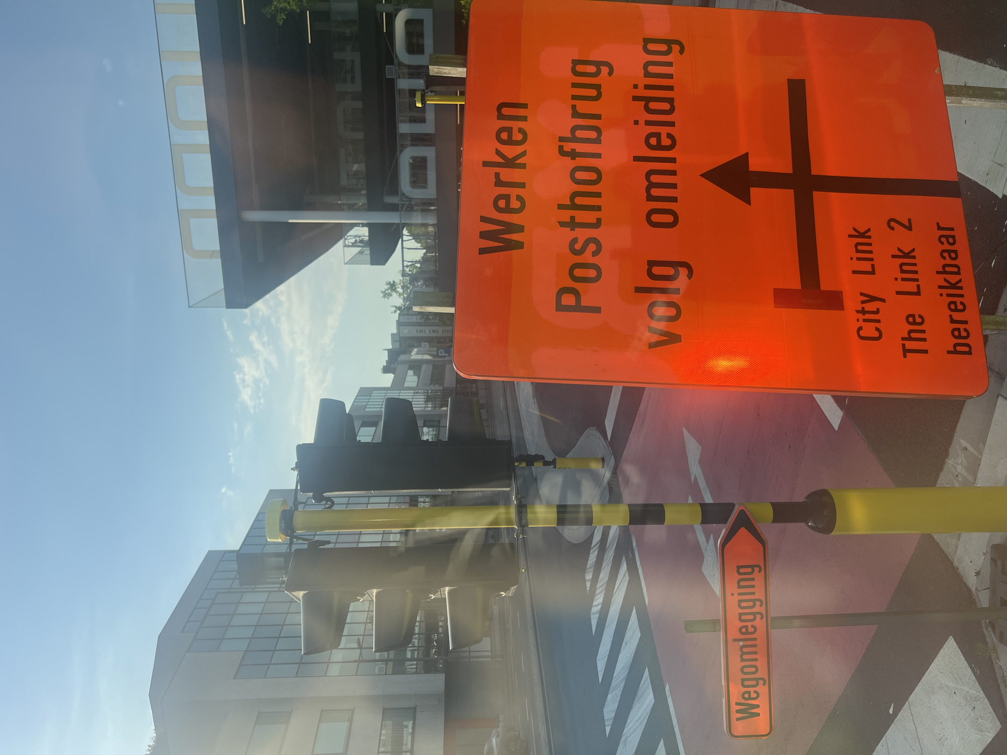 The image depicts a street scene in Antwerp, Belgium, dominated by an orange road construction sign. The sign reads "Werken Posthofbrug volg omleiding," meaning "Work Posthof Bridge follow detour." Below this, a diagram indicates a detour route with arrows showing a straight path, a right turn, and a left turn. Further text on the sign states "City Link The Link 2 bereikbaar," indicating that City Link and The Link 2 are accessible.
In the foreground, a yellow traffic signal pole with black bands stands prominently, partially obscured by sunlight glare. A smaller orange directional sign with the text "Wegomlegging" (detour) points to the right. The ground features a red-marked pedestrian crossing and a bike lane.
In the background, modern buildings with glass facades and a structure with large yellow lettering are visible under a clear blue sky with scattered clouds. The overall scene suggests active road construction and traffic management in an urban setting. There are no people or vehicles clearly visible in the image.