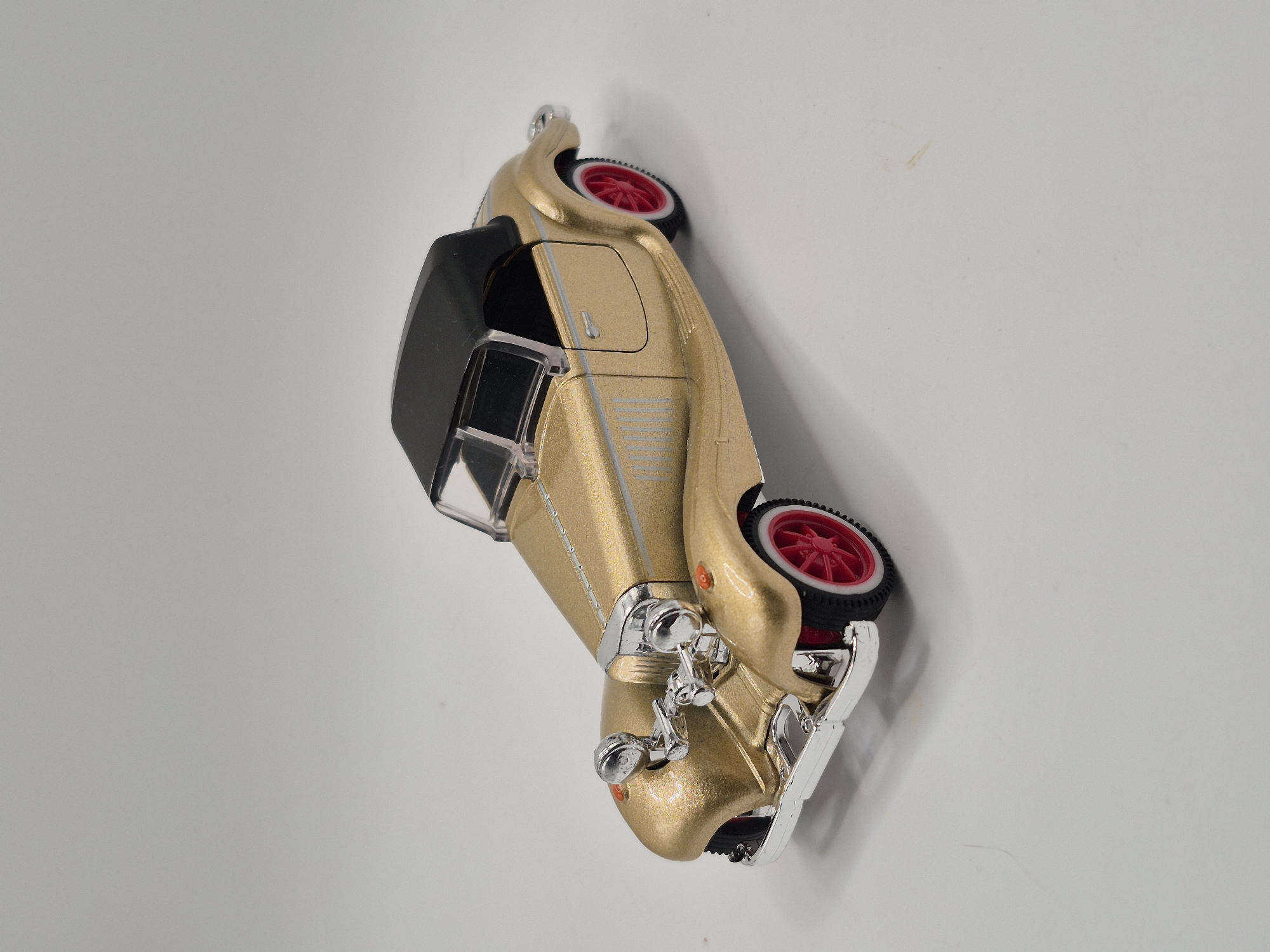 A gold vintage-style toy convertible car with a black soft top and red wheels sits on a plain white surface. The car has chrome accents on the grille, headlights, bumpers, and door handles. Vertical silver stripes are present on the car's side panels. The image is a studio shot with artificial lighting, casting a subtle shadow to the lower left of the car. The background is a seamless, neutral gray. There are no people, actions, or interactions depicted. This image is not related to Schwäbisch Gmünd, Germany.