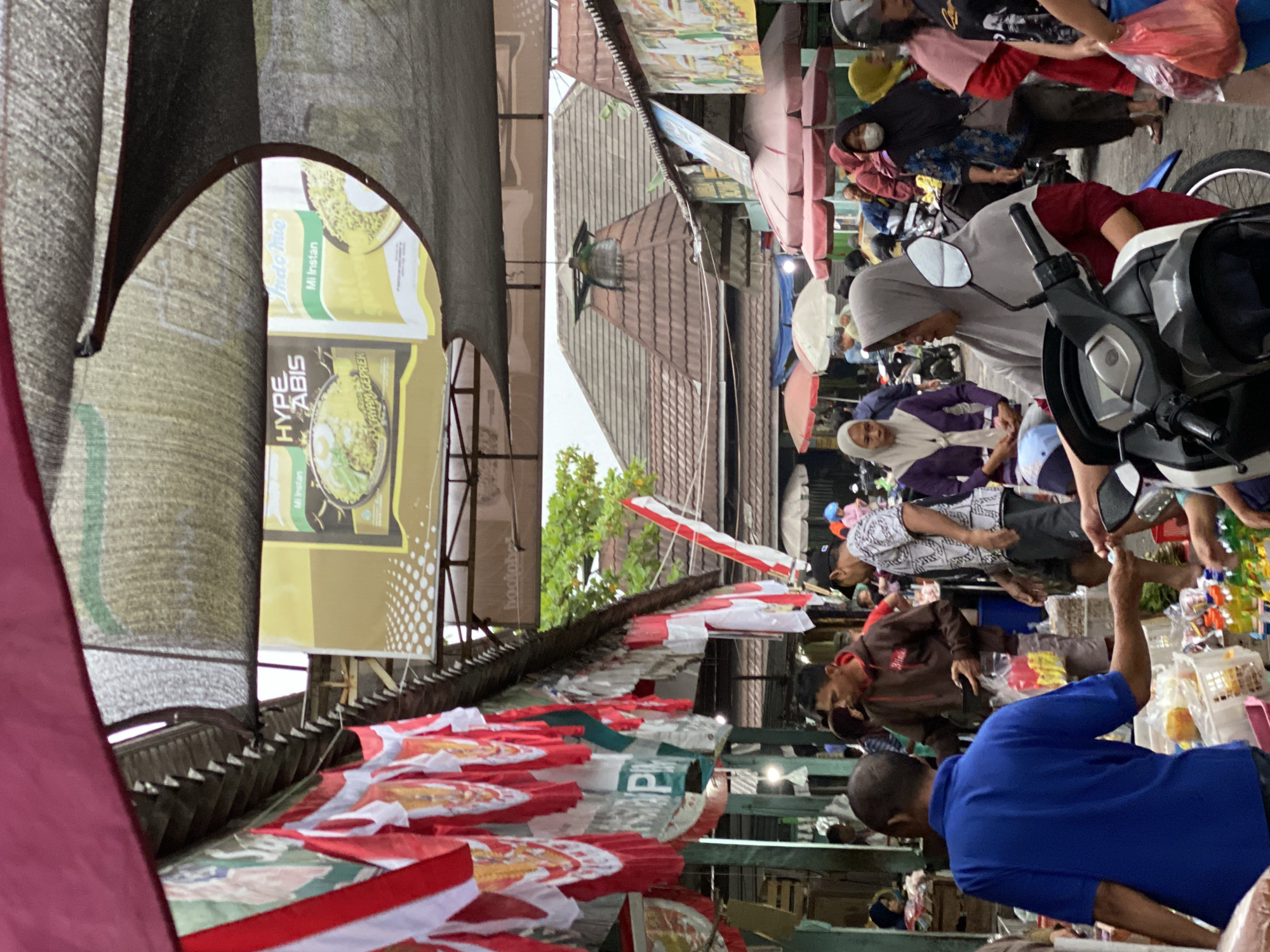 The image depicts a bustling market scene, likely in Ambarketawang, Indonesia, characterized by its vibrant atmosphere and the presence of numerous vendors and shoppers. Red and white Indonesian flags are prominently displayed, suggesting a patriotic occasion or general nationalistic pride. The market appears to be covered, with tarps and awnings providing shade, and hints of traditional architecture can be seen in the background.
In the foreground, a man in a blue shirt is interacting with someone off-camera, possibly engaged in a transaction. His arm is extended, and he appears to be handing something over. Behind him, a man in a brown hoodie is holding a wallet and looking down, while another man in a patterned shirt stands nearby. Several people are wearing masks, indicating adherence to health protocols. A variety of goods are visible, including bottled drinks and items in baskets. The presence of motorcycles suggests that transportation is a common mode of arrival and departure for market-goers. The overall impression is one of lively commerce and community activity within a traditional Indonesian market setting.