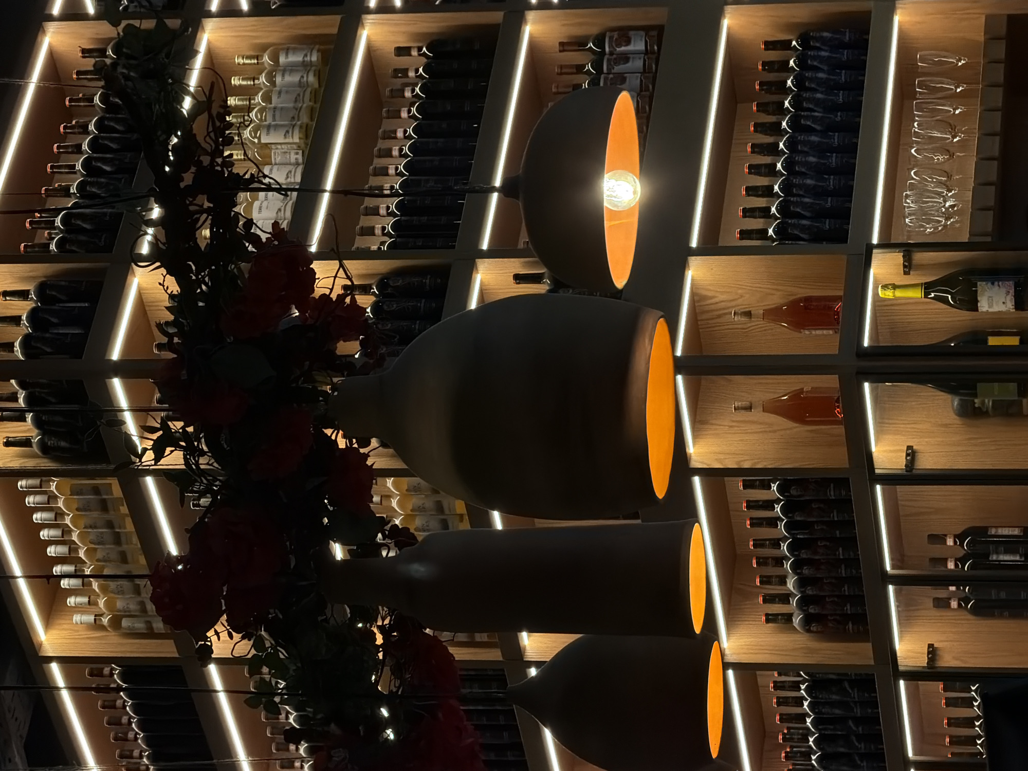 The image depicts the interior of a wine cellar or a restaurant with a wine display. The shelves are stocked with numerous bottles of wine and some empty wine glasses. Three pendant lights with orange interiors are hanging from the ceiling, one of which has a visible light bulb. Decorative red flowers and green foliage are also present, hanging above the wine bottles. The lighting is dim and atmospheric, with LED strips illuminating the shelves. The scene does not contain any people. The image does not provide any specific information that would allow for the determination of its location in the city of Perm, Russia.