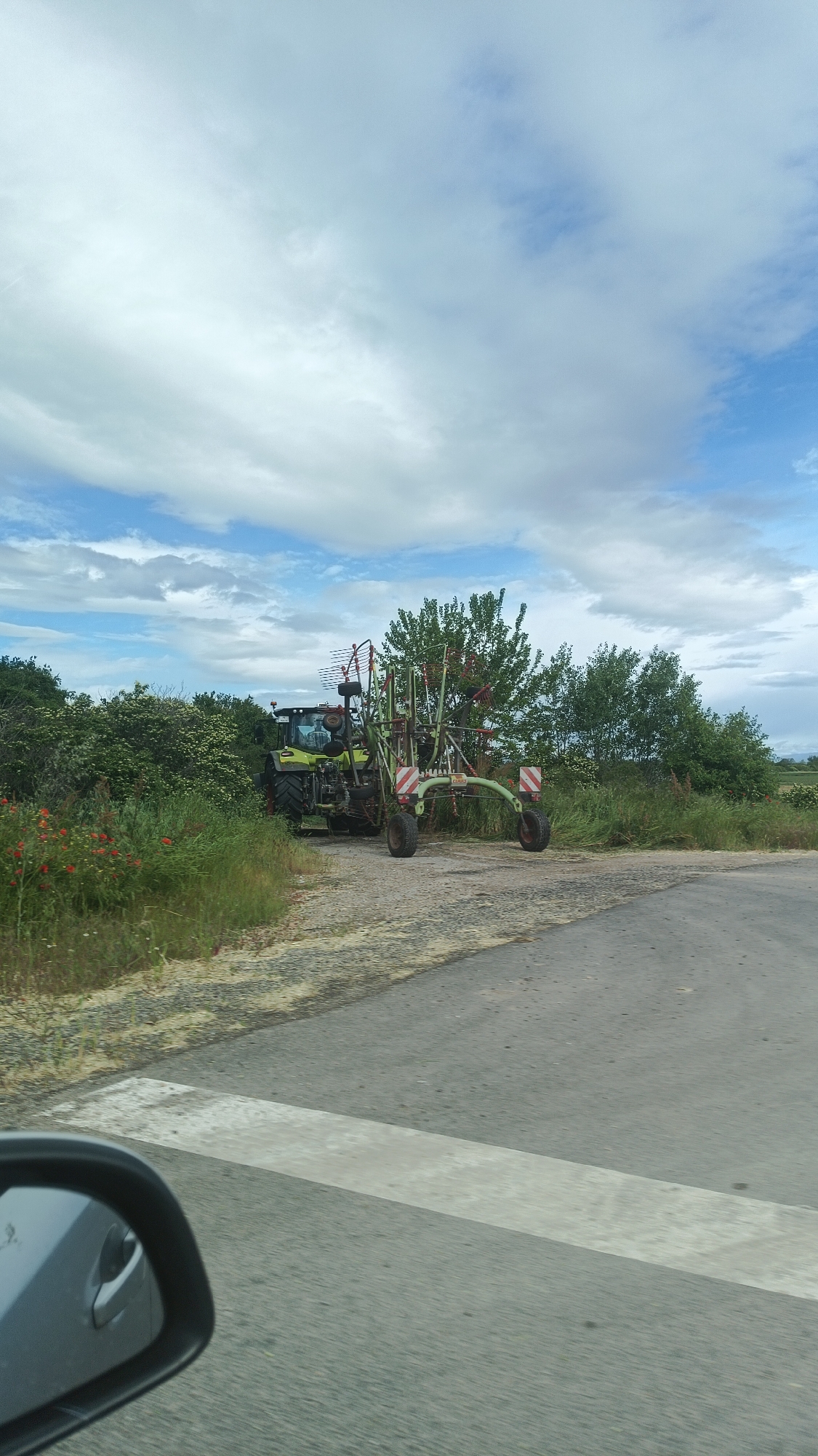 The image shows a green CLAAS tractor with an attached agricultural implement stopped on a paved road at a crossroads. The location appears to be rural, with vegetation, including some red poppies, lining the roadside. A car mirror is visible in the lower-left corner of the image, suggesting the photo was taken from inside a vehicle. The sky is partly cloudy. There are no people visible outside the tractor. The road appears to be a narrow country lane, possibly in Milevo, Bulgaria.