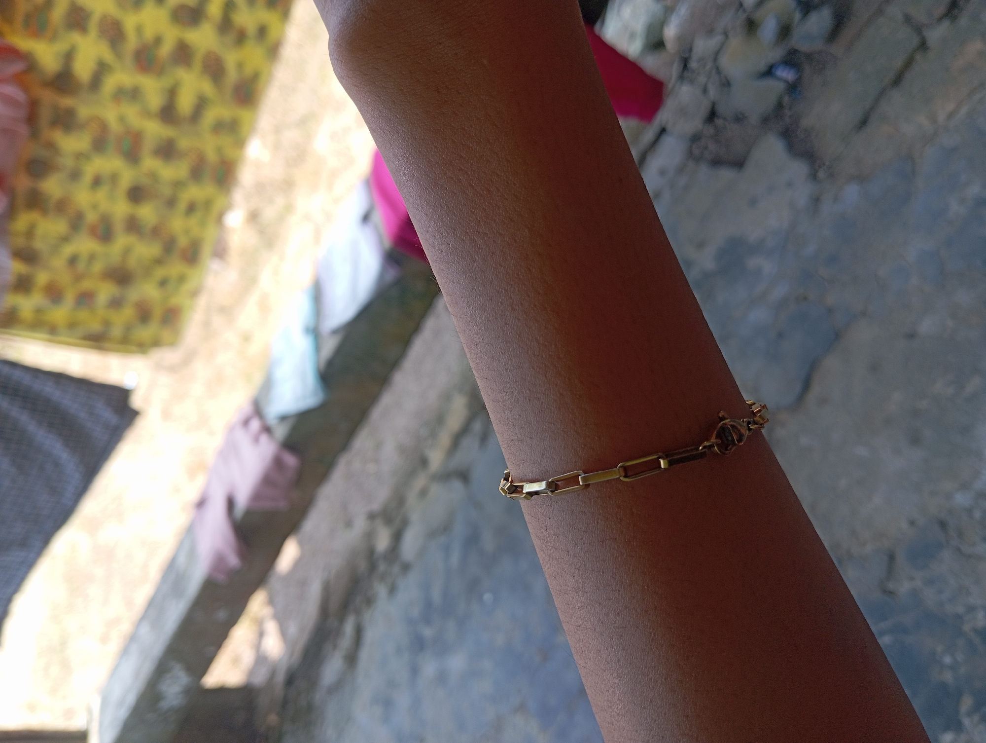 A person's forearm, adorned with a gold chain bracelet, is positioned diagonally across the frame. The skin is a light brown with visible pores. The bracelet, made of interlocking rectangular links, features a small charm. In the background, a concrete ledge with a pink garment draped over it is visible, along with a blurry yellow patterned fabric. The ground appears to be a rough, textured surface. The scene is outdoors, possibly in a courtyard or a casual outdoor setting. The overall impression is one of casual observation.