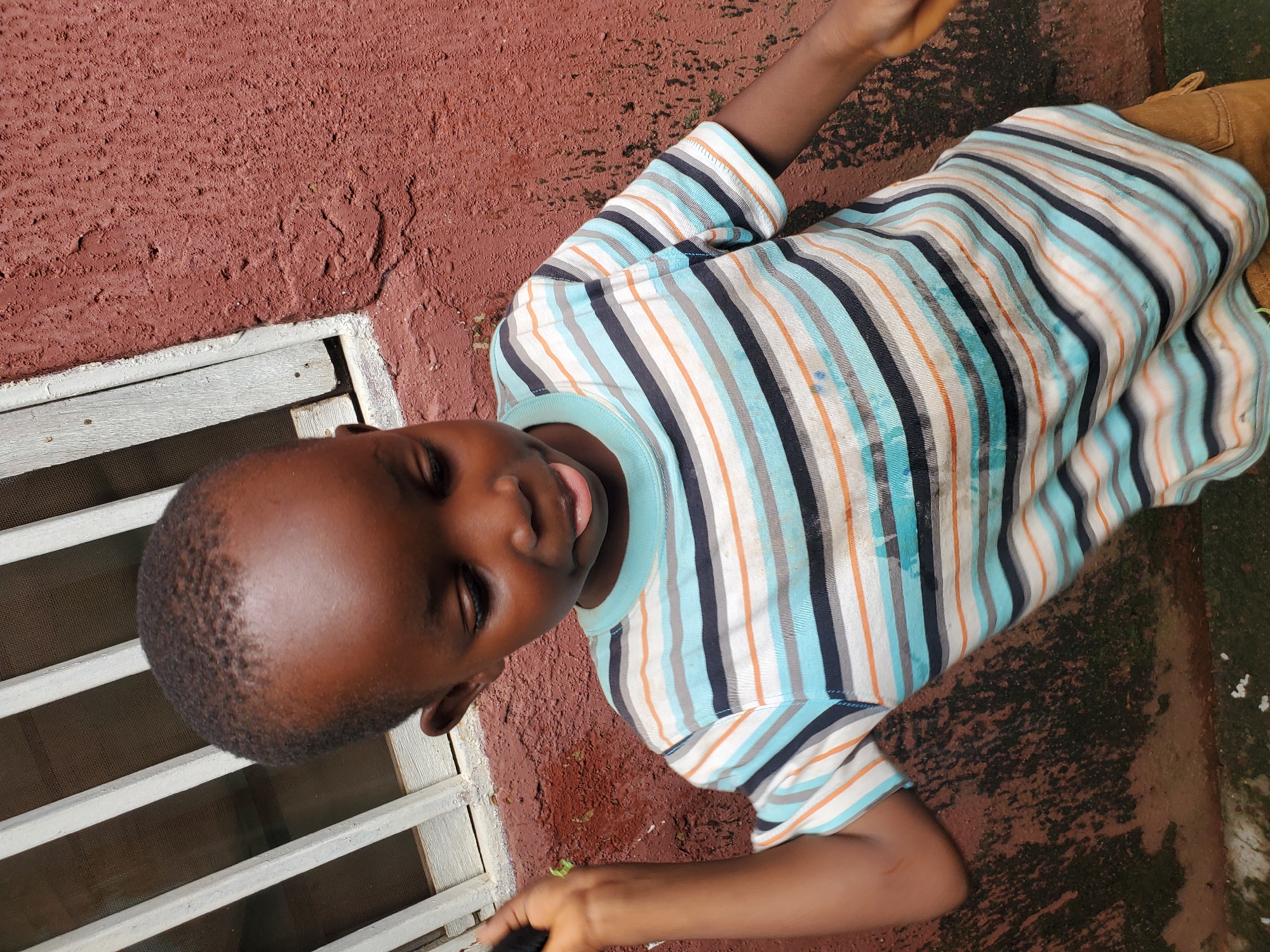 The image shows a young Black child lying down, with their head tilted upwards. The child is wearing a striped t-shirt with colors of blue, white, orange, and black. Their eyes are closed, and their tongue is slightly sticking out, suggesting a relaxed or playful mood. The background consists of a textured reddish-brown wall, a white barred window with a screen, and some dark green, mossy ground. The lighting suggests it might be daytime, possibly outdoors given the textures and colors of the environment. The overall scene appears to be in a somewhat rustic or utilitarian setting. There is no visible text in the image. The context of Jos, Nigeria, implies an outdoor setting that is likely warm.