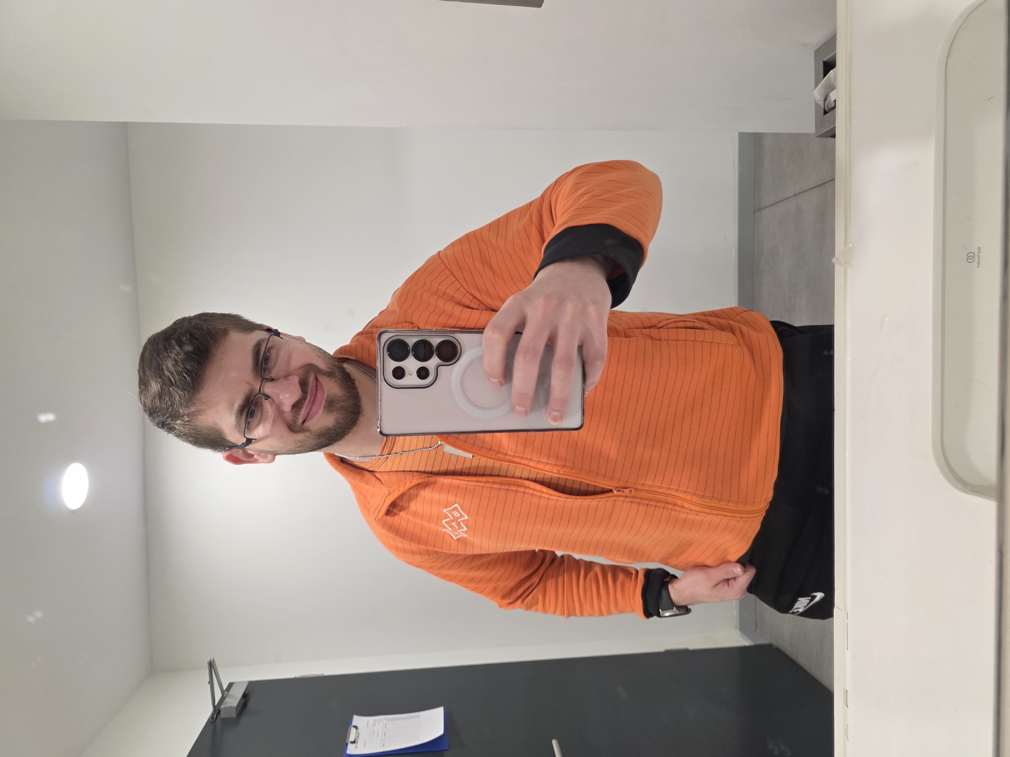 A man with glasses, a beard, and an orange jacket takes a selfie in a bathroom. He is holding a phone with a white case. He is wearing black athletic shorts. The bathroom has a light wall, a dark gray baseboard, a white sink and countertop, and a white toilet. There is a paper on a clipboard attached to the wall. The location in Plovdiv, Bulgaria is not evident from the picture.