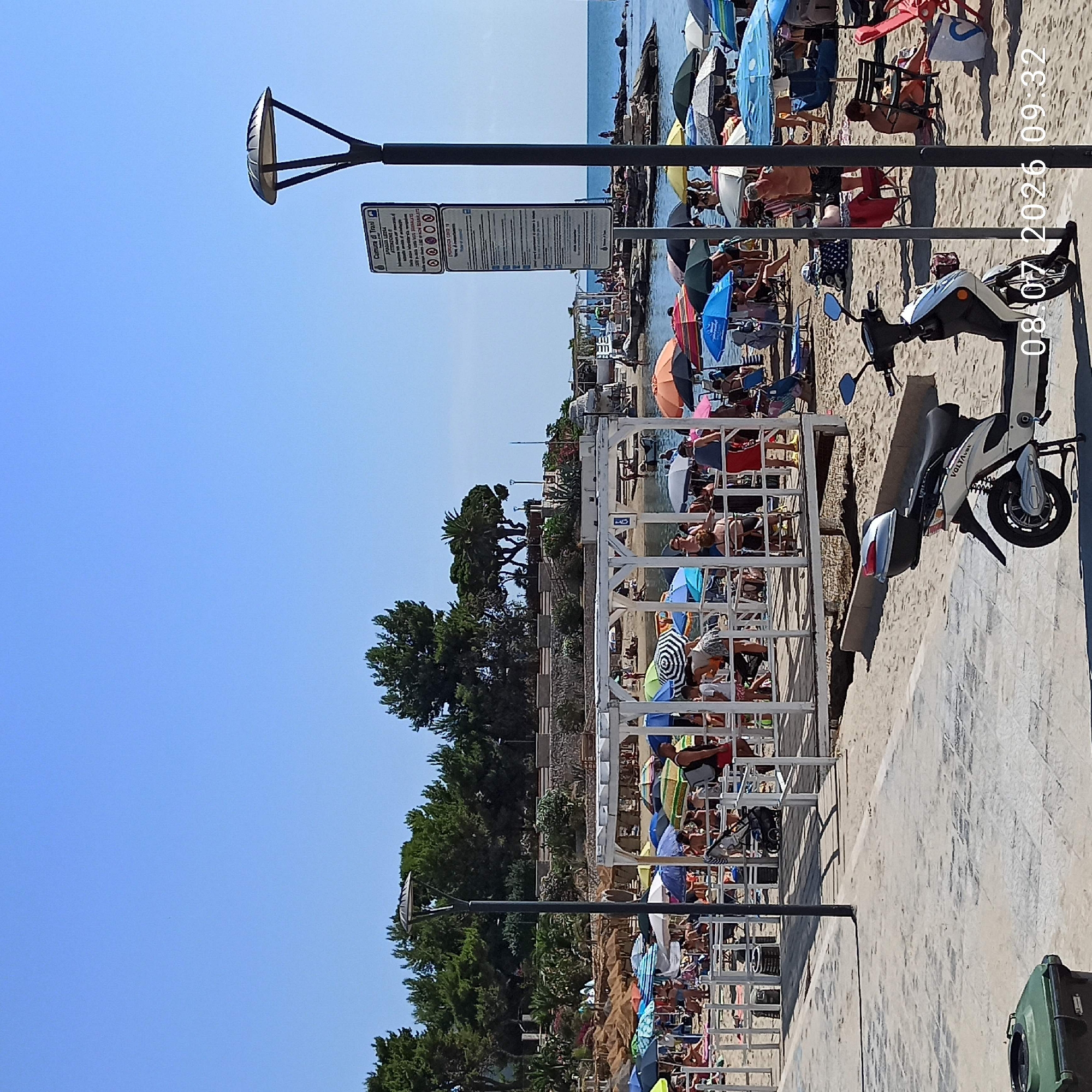 The image depicts a crowded beach scene in Trani, Italy. The beach is lined with numerous colorful umbrellas providing shade for beachgoers. People are scattered across the sand, some lounging under umbrellas, others walking or sitting. A white wooden structure, possibly a pavilion or a series of cabanas, is prominent in the foreground. In the lower right, a white scooter is parked. A lamppost stands to the right of the image, and a sign with Italian text is attached to its pole. The sea is visible in the background, with a clear blue sky above. The overall impression is a busy, sunny day at a popular beach.