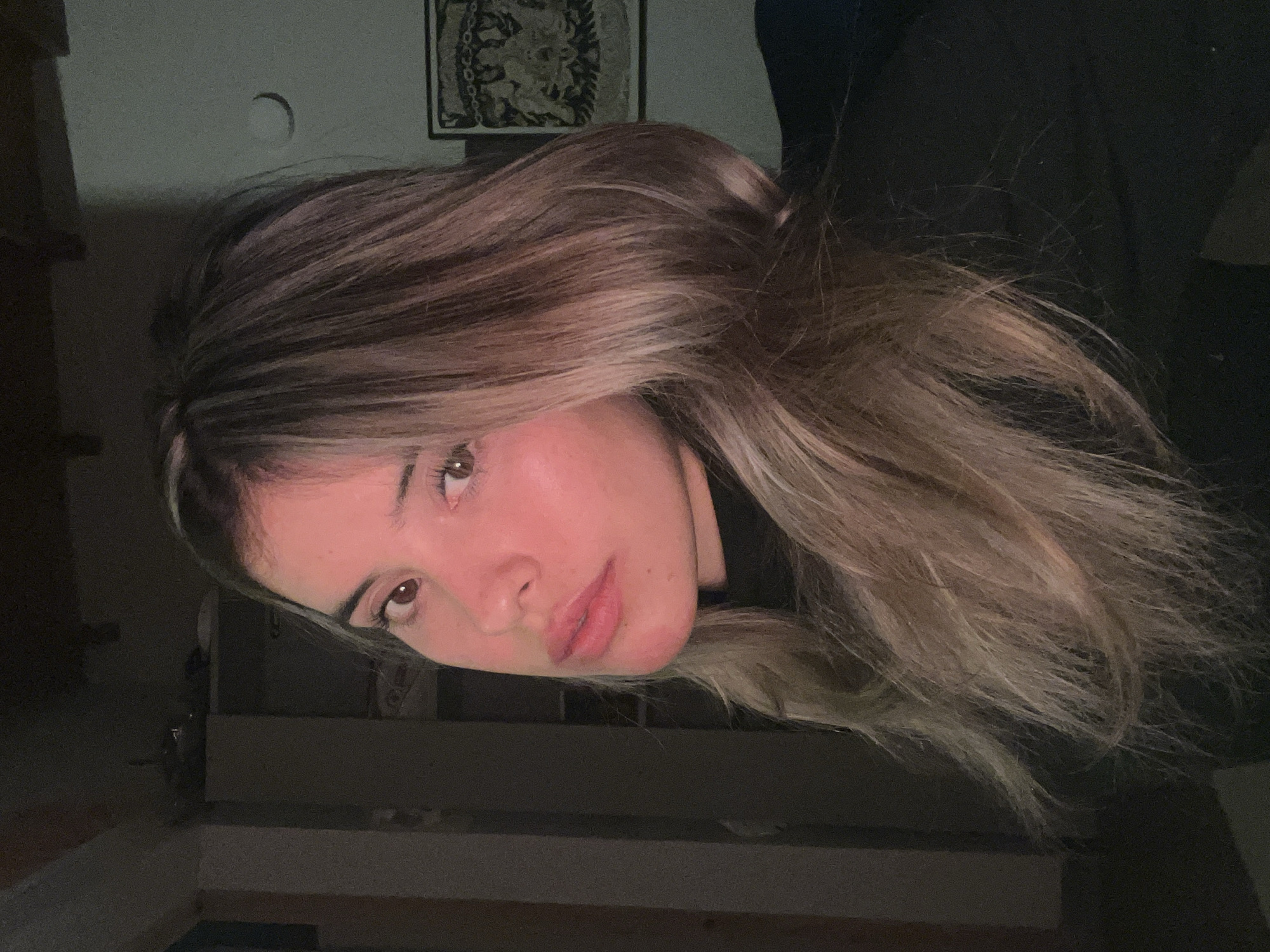 The image is a close-up, waist-up portrait of a young woman with blonde and brown ombre hair, wearing a black turtleneck. She is looking off to the left of the frame, with her face illuminated by warm lighting, casting a pinkish glow on her skin. Her lips are parted slightly, and her eyes are wide open. In the background, a dark wall is visible, with a framed tapestry or artwork on the right side and a circular object that could be a light fixture or a decoration on the wall. The overall environment appears to be indoors, likely a dimly lit room. There is no discernible information within the image to suggest the location is Sumalao, Argentina, or any other specific city.