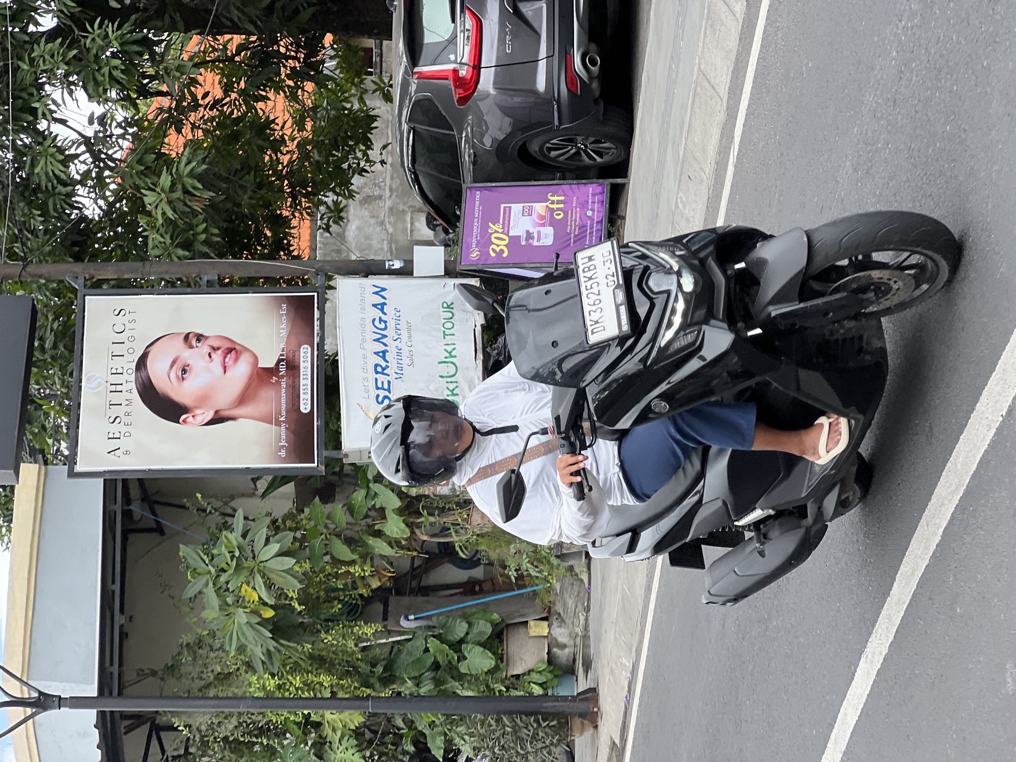 A medium shot captures a person riding a black scooter down a paved road in Denpasar, Indonesia. The rider, wearing a white long-sleeved shirt, blue pants, and a black helmet with a clear visor, is positioned in the center of the frame. A tan strap is visible across the rider's chest. The scooter has a clear windshield and a license plate displaying "DK 3625 KBW".
To the left of the rider, a tall black lamppost stands beside a building with a light-colored facade. Lush green foliage is abundant, with plants in pots and growing wild.
To the right of the rider, a gray SUV is parked. Next to the SUV, a purple advertisement displays "30% off" and "REJUVENIQUE AESTHETICS". Further down the street, another advertisement reads "AESTHETICS & DERMATOLOGIST". The background is filled with trees and an orange-tiled roof. The road is marked with white lines. The lighting suggests it is daytime, possibly overcast or late afternoon.