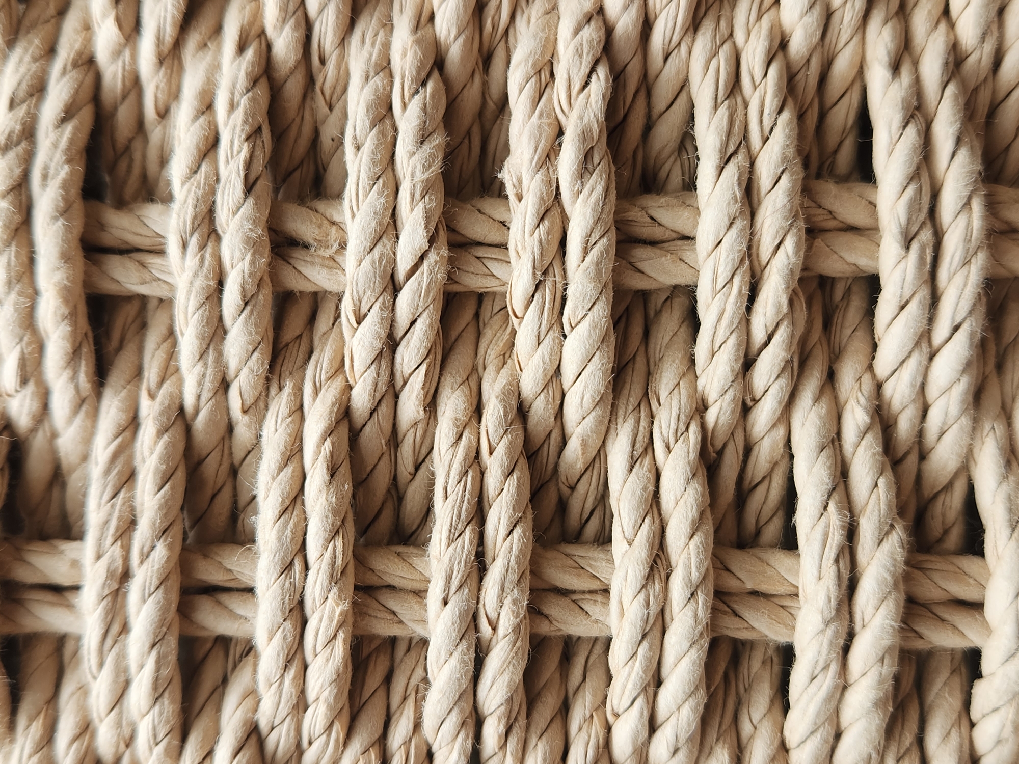 A close-up, textured shot of a woven basket. The basket appears to be made of thick, natural fiber ropes, possibly jute or sisal, woven in a crisscross pattern. The ropes are a light, earthy beige color and have a slightly fuzzy or fibrous texture. The weaving is tight and even, creating a distinct grid-like appearance with some depth where the horizontal and vertical ropes intersect. There are no people, animals, or discernible surroundings visible. The image is focused entirely on the materiality and structure of the woven basket.