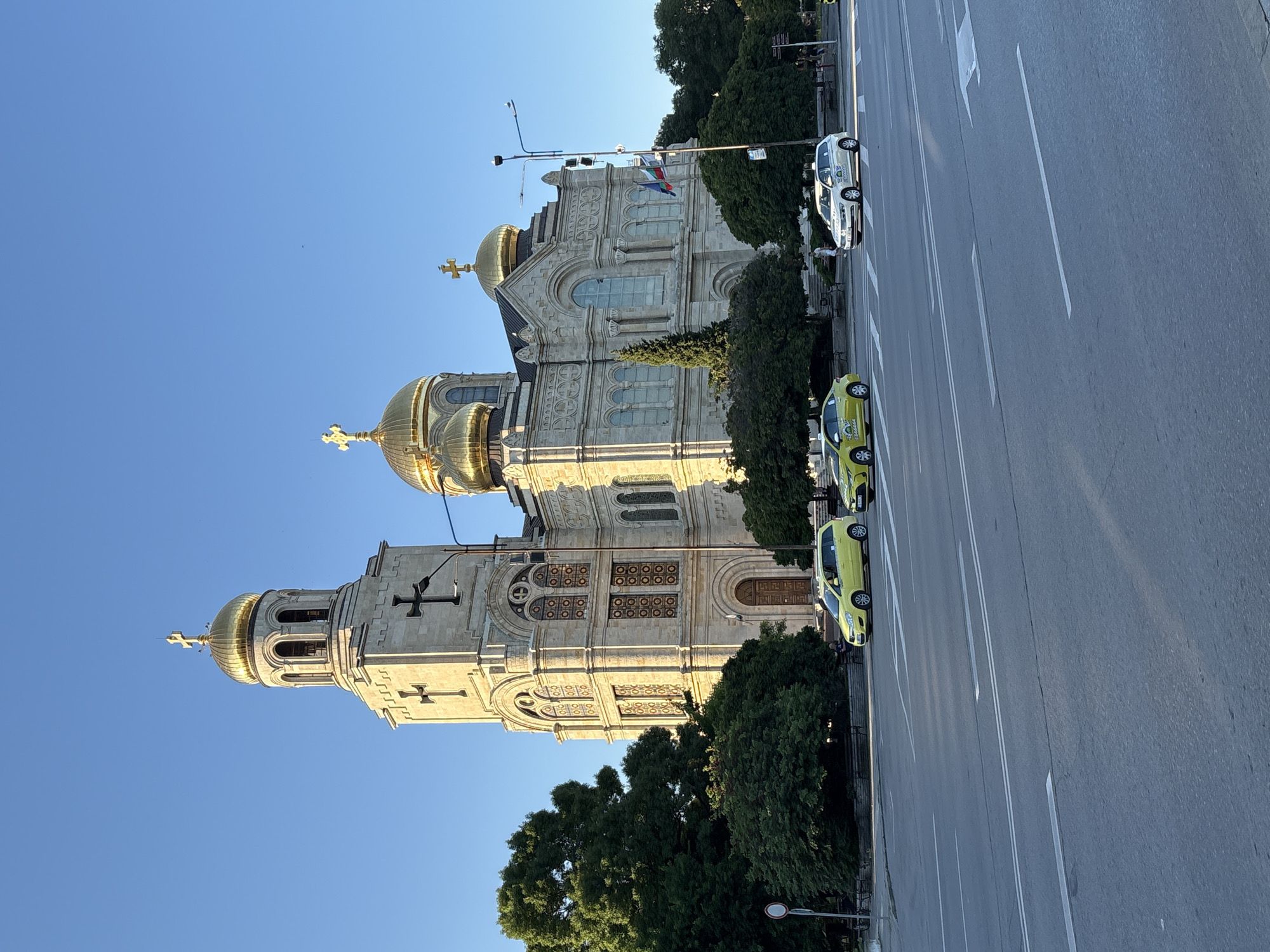 The image depicts the Cathedral of the Assumption of the Virgin in Varna, Bulgaria, on a clear day with a bright blue sky. The grand stone cathedral features multiple golden domes and a tall bell tower, both adorned with crosses. The facade is intricately detailed with arched windows and decorative elements.
In the foreground, a multi-lane road runs in front of the cathedral. Several vehicles are visible on the road, including three yellow taxis and one white car that appears to be a taxi as well. The yellow taxis are driving on the right side of the road, while the white car is in the left lane, also moving to the right. A few pedestrians are walking on the sidewalk in front of the cathedral.
Lush green trees partially obscure the lower sections of the cathedral and extend to the sides of the image. Flags of Bulgaria are visible on a flagpole near the cathedral. The overall scene is that of a public space, likely a city square or intersection, with the prominent cathedral serving as the central landmark. The sunlight casts a warm glow on the building, suggesting it is late afternoon or early morning.