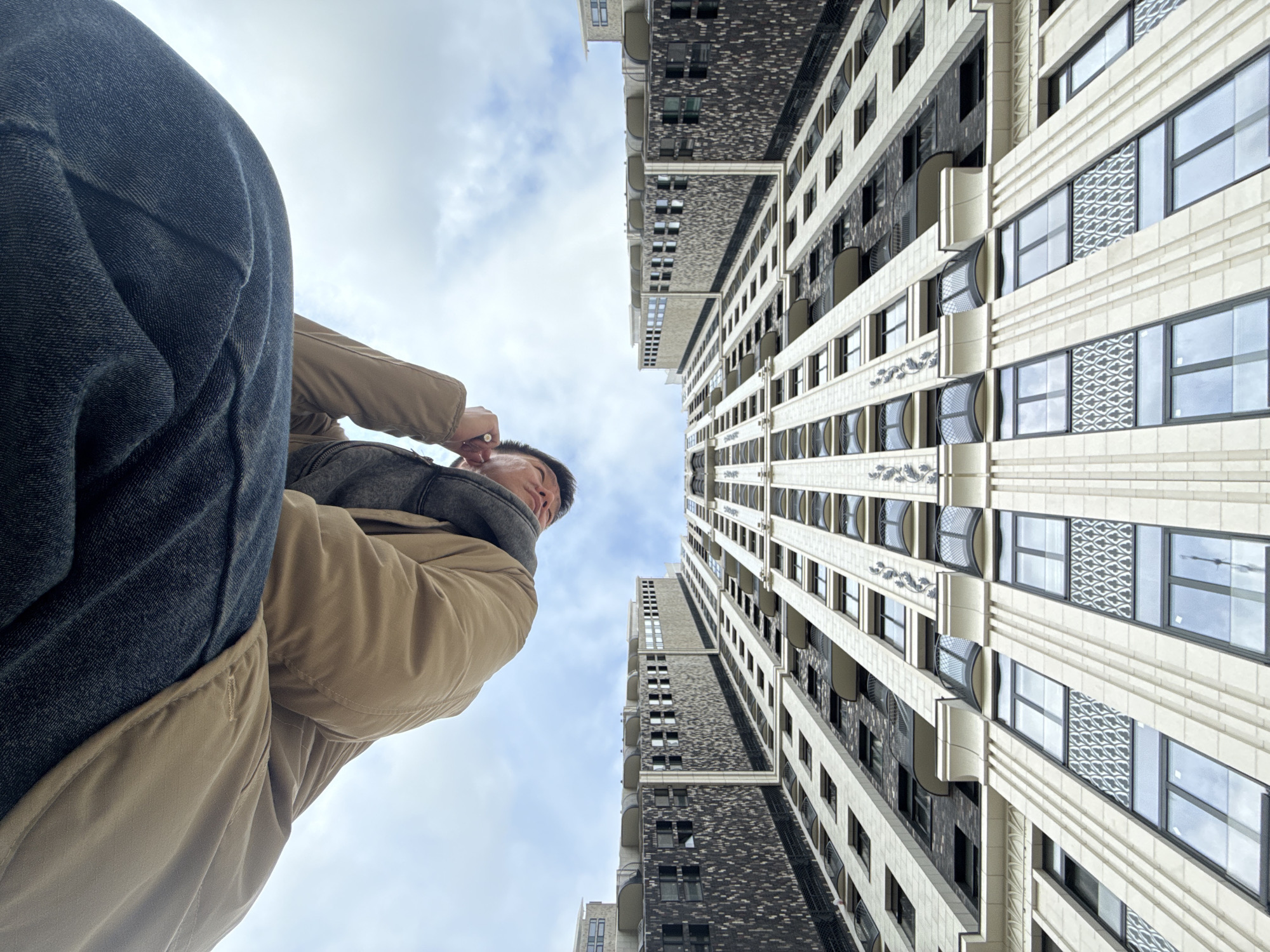 A low-angle shot shows a person looking up at two high-rise buildings in Khimki, Russia. The person is wearing dark jeans, a tan jacket, and a gray scarf. Their head is tilted back, and their hand is raised towards their face. The buildings are tall and narrow, with many windows. The building on the right is clad in light-colored stone with art deco details, while the building on the left is clad in a darker material with a more modern design. The sky is partly cloudy.