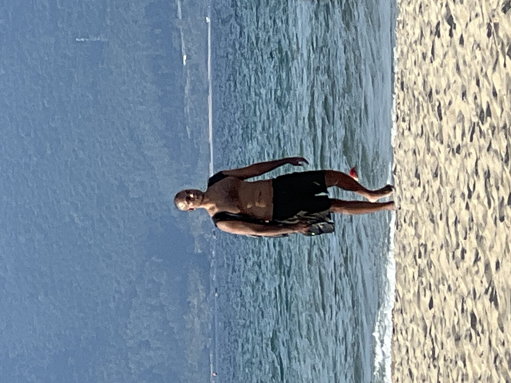 A bald, fair-skinned man with dark hair on his chest walks on a sandy beach toward the viewer. He is wearing black swim shorts and a black backpack with a light-colored object tucked into it. The man is facing forward with his left arm slightly bent. The ocean, with small waves, is behind him, and a forested mountainside is in the background. The scene is likely located in Đà Nẵng, Vietnam, known for its beaches and coastal mountains.