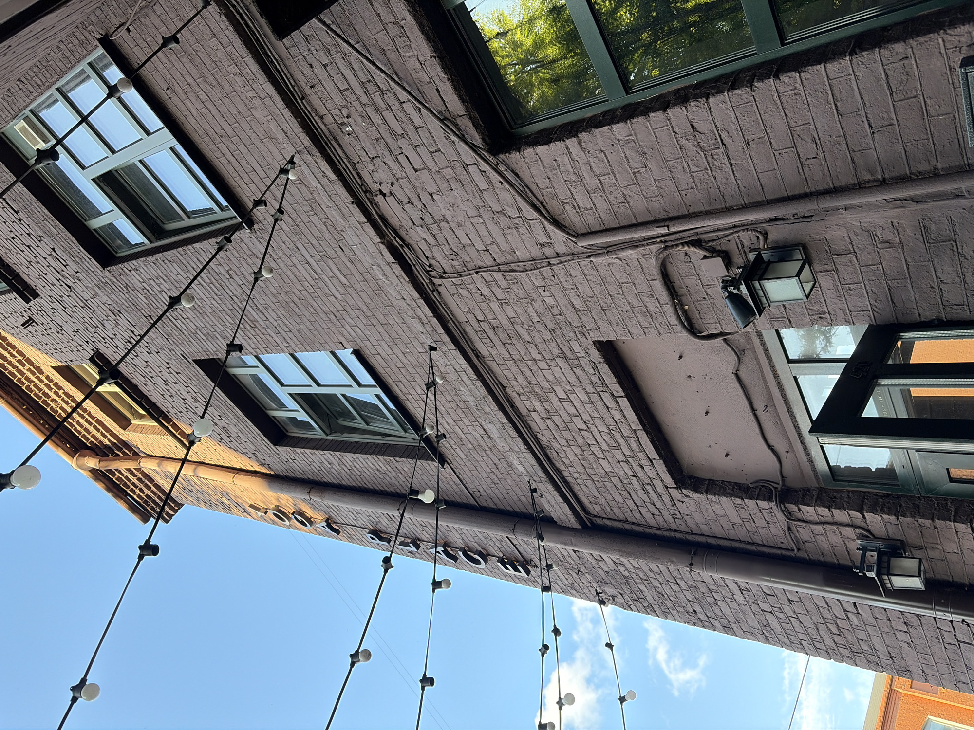 The image shows an exterior view of a brick building with an urban setting. The building is painted a dark reddish-brown color and features several windows with green frames. String lights with visible bulbs are suspended across the building and the sky, which is a clear blue with a few wispy clouds. A vertical pipe runs down the side of the building. There are also outdoor lights mounted on the wall. In the background, another brick building with orange brickwork is partially visible. The angle of the shot is looking upwards. Text is visible vertically on the building, appearing to spell out "PIZZA CLUB". There are no people or animals in the image. The overall impression is that of an outdoor seating area for a restaurant or bar. It is not possible to determine the specific location in Minsk, Belarus from the provided image.