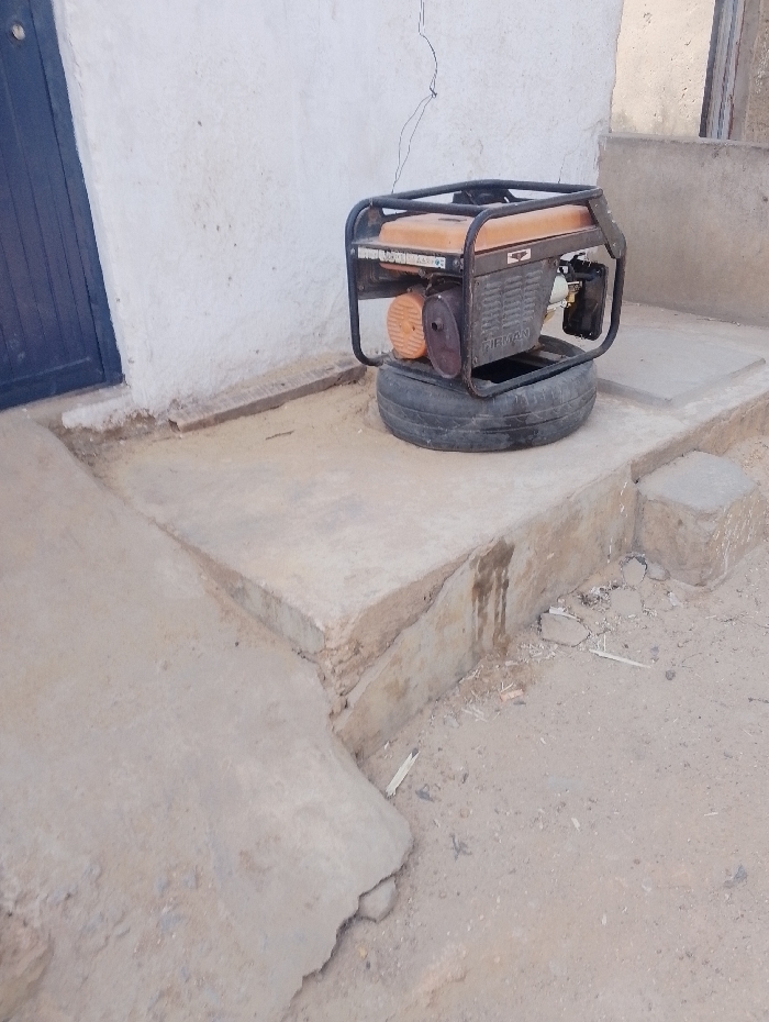 A Firman portable generator, featuring an orange fuel tank and a black metal frame, rests on a black rubber tire. The tire is positioned on a raised, multi-level concrete platform. The generator's engine block is grey, with an orange fan housing visible on its left side. Dark, vertical stains are present on the side of the concrete platform below the generator. Behind the platform, a textured white wall stands, partially revealing a dark blue door on its left side. A thin, dark wire or cable hangs vertically along the white wall. The ground in the foreground and to the right of the platform consists of dry, light-brown sandy soil, scattered with small pieces of light-colored debris. No individuals, actions, or interactions are present in this static scene, located in Dorayi, Nigeria.