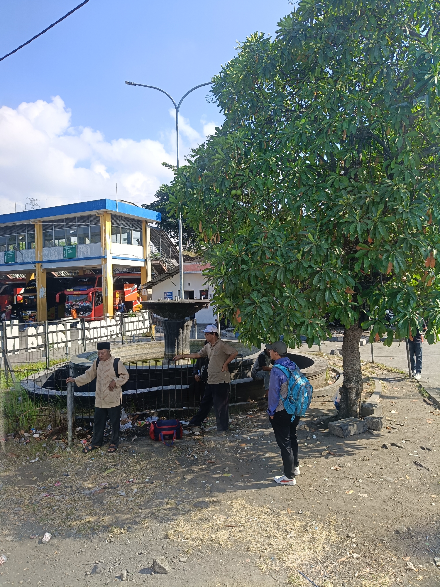 The image captures a daytime scene at a bus station in Kedungrejo, Indonesia. In the foreground, three men are standing near a low, circular, stone structure, possibly a fountain or planter, which is surrounded by a dark metal fence. One man, wearing a light-colored traditional garment and a black cap, leans on a pole. Another man in a tan shirt and white cap points towards the stone structure. A third man, wearing a blue jacket and a backpack, stands facing away from the camera. The ground is a mixture of dirt and gravel, with scattered debris and dry vegetation.
Behind the men and the stone structure is a large tree with dense green foliage. In the background, a two-story building with a blue roof and yellow walls houses several buses, indicating it is the bus terminal. The sky above is clear blue with scattered white clouds. A street lamp on a tall pole is visible to the left of the tree. The overall impression is of a functional, somewhat weathered, outdoor transportation hub on a sunny day.