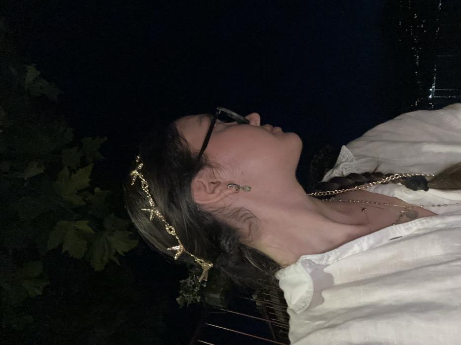 A low-angle shot shows a woman gazing upwards in the dark. She wears glasses and a star-shaped hair accessory, along with delicate earrings. Multiple necklaces adorn her neck, layered over a white collared shirt. A portion of her braided hair is visible. Some foliage is visible in the lower left corner, adding to the outdoor setting. The scene is dimly lit, emphasizing the upward gaze.