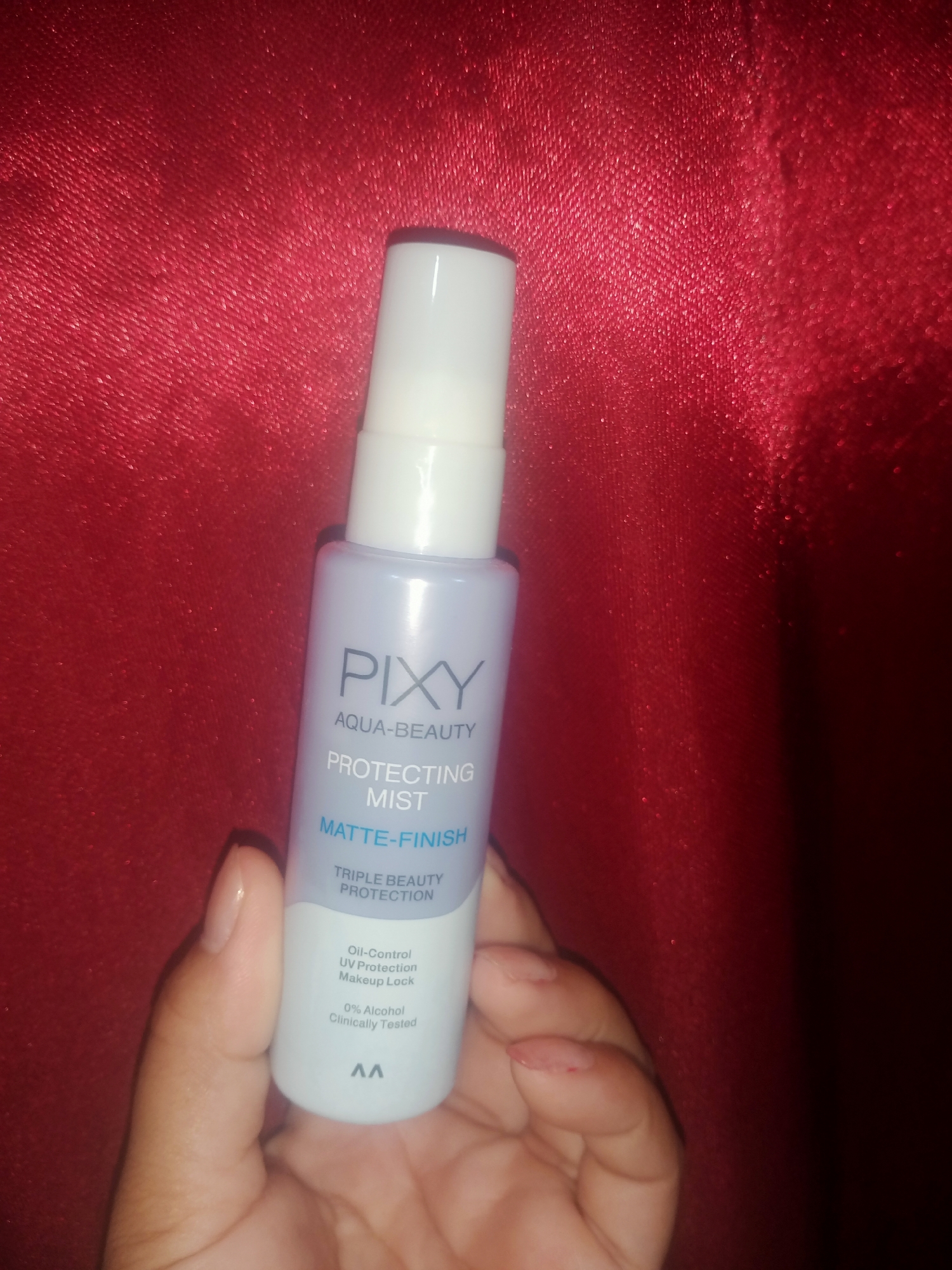 A light-purple, translucent plastic bottle of PIXY AQUA-BEAUTY PROTECTING MIST MATTE-FINISH is held in a person's left hand, in front of a red, textured fabric background. The bottle has a white cap and a label with blue text. The text on the label reads "PIXY", "AQUA-BEAUTY", "PROTECTING MIST", "MATTE-FINISH", "TRIPLE BEAUTY PROTECTION", "Oil-Control UV Protection Makeup Lock", "0% Alcohol Clinically Tested", and a logo that appears to be two connected 'A's. The fabric background appears to be a satin or silk material with a crinkled texture. The scene is indoors, likely in a domestic setting. There are no other people or significant objects visible in the image. The lighting appears to be artificial, casting a slight shadow to the right of the bottle. The image is taken from a slightly elevated angle, looking down at the bottle and hand. The location of Nusa Agung, Indonesia, is not discernible from the image.