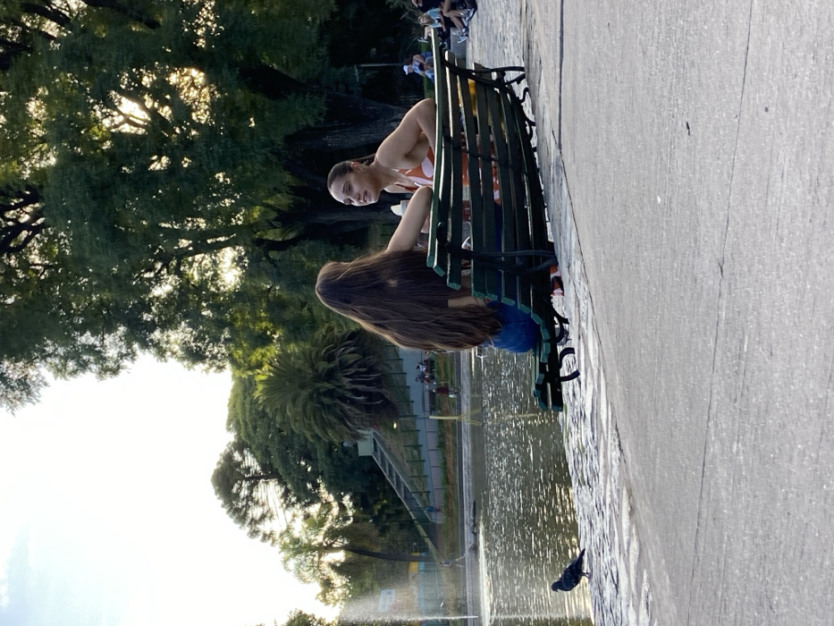 The image depicts an outdoor scene at Rosedal, located within the Bosques de Palermo in Buenos Aires, Argentina, during what appears to be late afternoon. Two adult females are seated on a dark green slatted park bench positioned parallel to the edge of a pond. The female on the left has long, dark brown hair, wears a dark-colored top and blue jeans, and is seen from behind. Her right arm is bent. The female on the right has lighter brown hair pulled back in a ponytail, wears an orange and white patterned top, and is facing the first female, smiling, and engaged in conversation. Her left arm rests on the back of the bench.
To the left of the bench, a dark-feathered pigeon stands on the paved bank immediately adjacent to the pond. Further left in the pond, a water fountain is active, spraying water upwards. The pond's surface exhibits ripples and reflections of light. The background is characterized by numerous large trees with lush green foliage, including deciduous trees and a palm tree, through which sunlight filters. In the distant background, a set of concrete stairs with a green railing ascends a gentle slope, and several indistinct figures are visible on grassy areas and by the water's edge. The foreground features a paved walkway, with a section of irregular, stone-like paving directly beside the pond and a smoother concrete path extending further away.