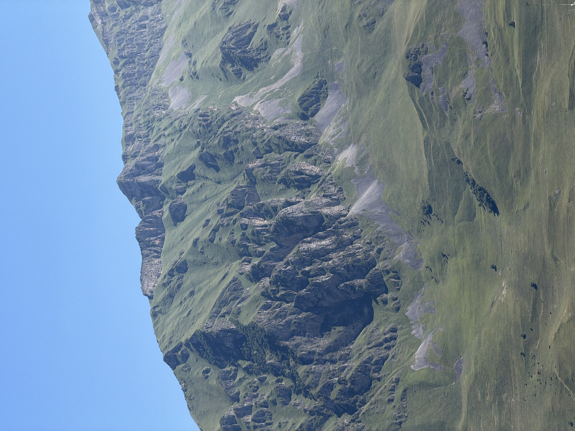 The image shows a mountainous landscape under a clear blue sky. The mountains are covered in green grass, with rocky outcrops and steep slopes. The terrain is rugged and uneven, with visible erosion patterns and small patches of trees on the lower slopes. The scene appears to be a natural, undeveloped area, likely in a rural or wilderness setting. No people, specific objects, or actions are discernible in the image. The location is identified as the city of Karmadon, Russia.