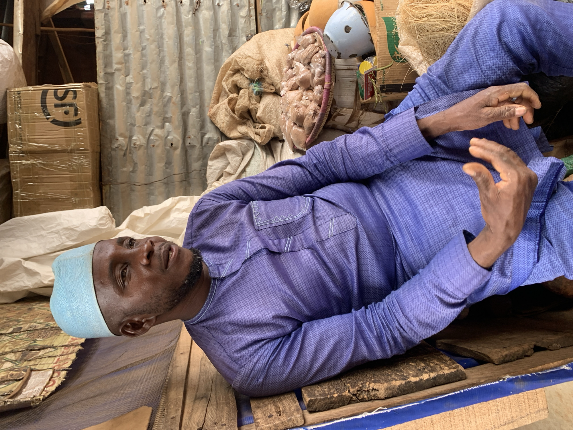 This media file shows a medium shot of a Black man wearing a blue skullcap and a purple patterned tunic, lying on his back. He appears to be in an outdoor or semi-outdoor setting, possibly a market or storage area, given the corrugated metal walls and stacks of goods in the background. To his left, there are cardboard boxes with the "SF" logo visible, wrapped in plastic. To his right, various items are stacked, including a woven basket filled with small, individually wrapped items, a large sack, and some straw-like material. The lighting suggests it could be daytime. The man's gaze is directed upwards and slightly to his left, and his expression appears neutral. His hands are resting on his chest and abdomen.
