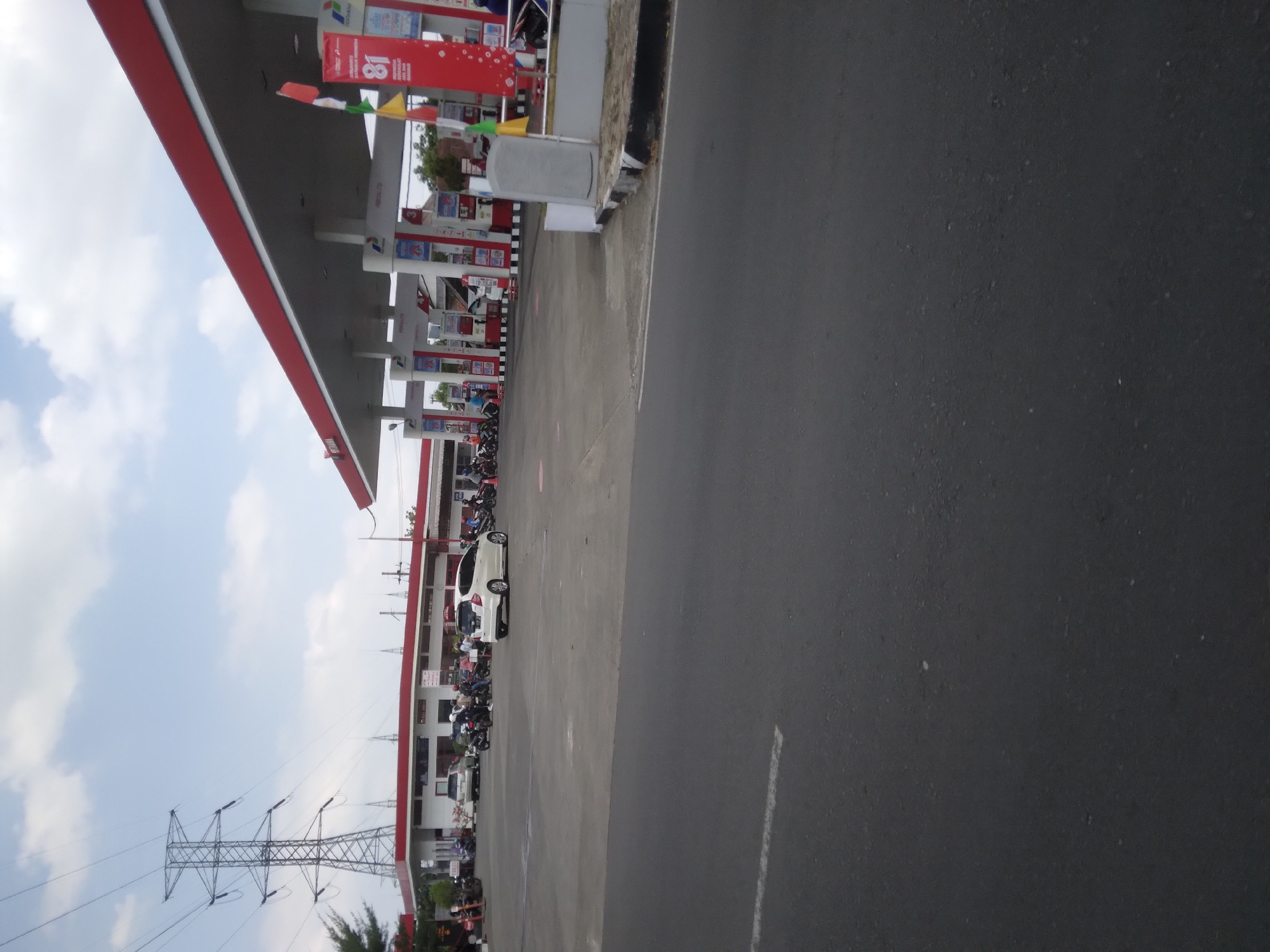 The image depicts a busy Pertamina gas station located in Purwokerto Kidul, Indonesia. The gas station features a red and white canopy covering multiple fuel pumps. Several vehicles, including cars and motorcycles, are present, with some actively refueling or parked. People are seen operating motorcycles and walking around the station. In the background, a large electrical tower stands tall against a cloudy sky. The foreground shows a paved road with white lane markings.