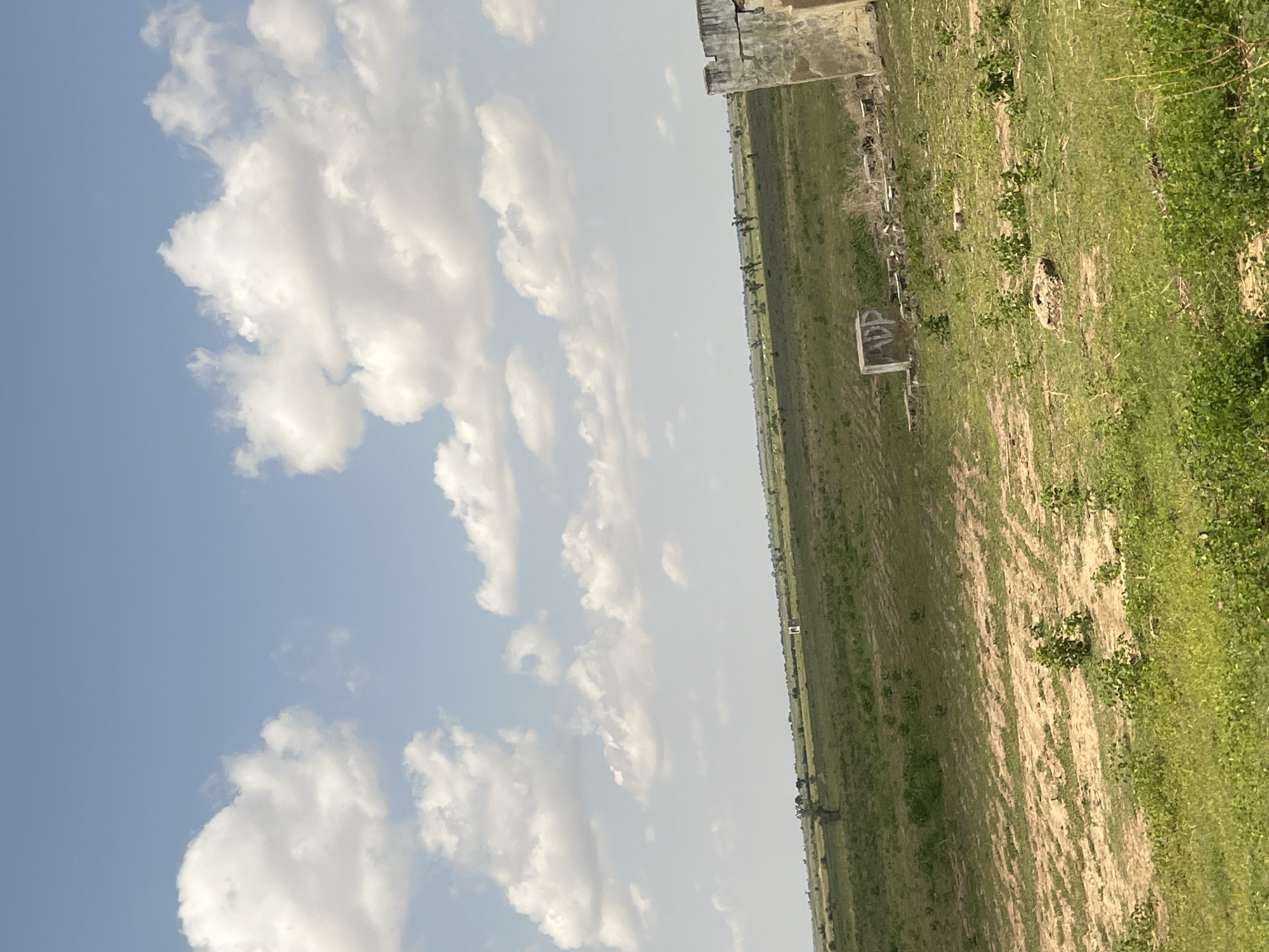 The image captures an expansive, gently sloping rural landscape under a partly cloudy sky, characteristic of an area in Kumo, Nigeria. The sky occupies approximately the upper two-thirds of the frame, displaying a light blue hue with numerous scattered white cumulus clouds of varying sizes. The lower third of the image is dominated by green vegetation, primarily grasses and small bushes, with several lighter-colored patches of exposed earth, possibly indicating tilled ground or dry areas.

In the immediate right foreground, a weathered, light-colored concrete or stone wall structure stands, appearing partially damaged with a jagged top edge and visible signs of deterioration. Further into the mid-ground, slightly left of center, a small, rectangular concrete block is visible on the ground, featuring dark, possibly graffiti-like markings on its side, which appear to include the letters "DP." Scattered debris, resembling broken concrete or bricks, lies on the ground near the foreground structures. Along the distant horizon, a small, light-colored, square building or structure is discernible, centrally positioned. Scattered trees and shrubs are visible across the middle and distant landscape. No individuals are present, and no direct human activity is depicted in the scene, which is illuminated by daylight.