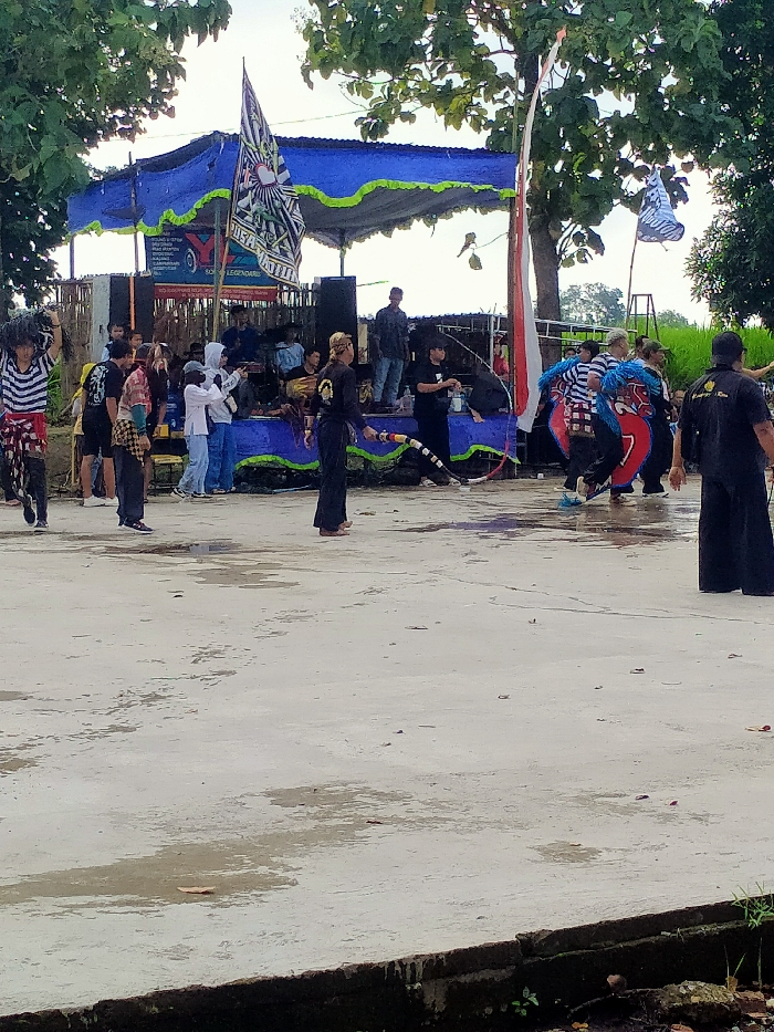 The image shows a performance taking place in Kecamatan Kedawung, Indonesia, at coordinates -7.519, 111.028. A group of people are gathered in front of a stage decorated with blue and white fabric and flags. The stage has a large banner with the word "LEGENDARY" printed on it. Some individuals are standing on the stage, while others are in front of it. There is a man in a black outfit holding a long, flexible rod. He is positioned in front of the stage, and is the focal point of the scene. Several other individuals are dancing in front of the stage. They are dressed in various colors, including black, white, and red. Some are holding flags and other objects, while others are simply moving their bodies to the music. The environment is outdoor, with a concrete surface and some trees in the background. It seems that it may have rained recently as there are puddles on the ground and it is possible that the event is taking place after a downpour. The overall atmosphere of the image is festive and celebratory.