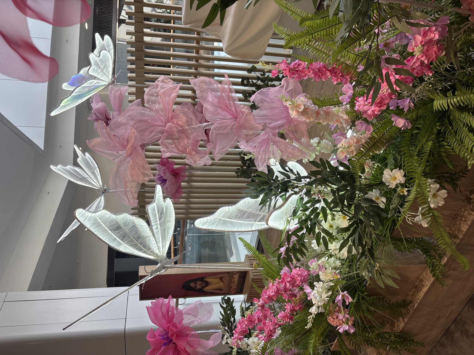 An indoor decorative display is observed within a modern commercial building, presumably a shopping mall, located in Izhevsk, Russia. The foreground features a dense, multi-tiered arrangement of real or artificial flora, predominantly green ferns, various pink flowers, and white blossoms, housed within wooden planters. Three large, illuminated butterfly sculptures with translucent, veined wings are positioned above the flora; one butterfly displays an iridescent blue-purple sheen. Several oversized, light pink fabric flowers, some embellished with small beads, are interspersed among the butterflies. To the left, a framed portrait of a young girl with dark hair, wearing a yellow top and with folded arms, is visible against a reddish-pink panel. The background reveals architectural elements including light-colored walls, a ceiling, glass railings suggesting an upper level, and a wooden slatted partition. A faint reflection of a human silhouette is visible in the background glass. The scene is brightly illuminated.