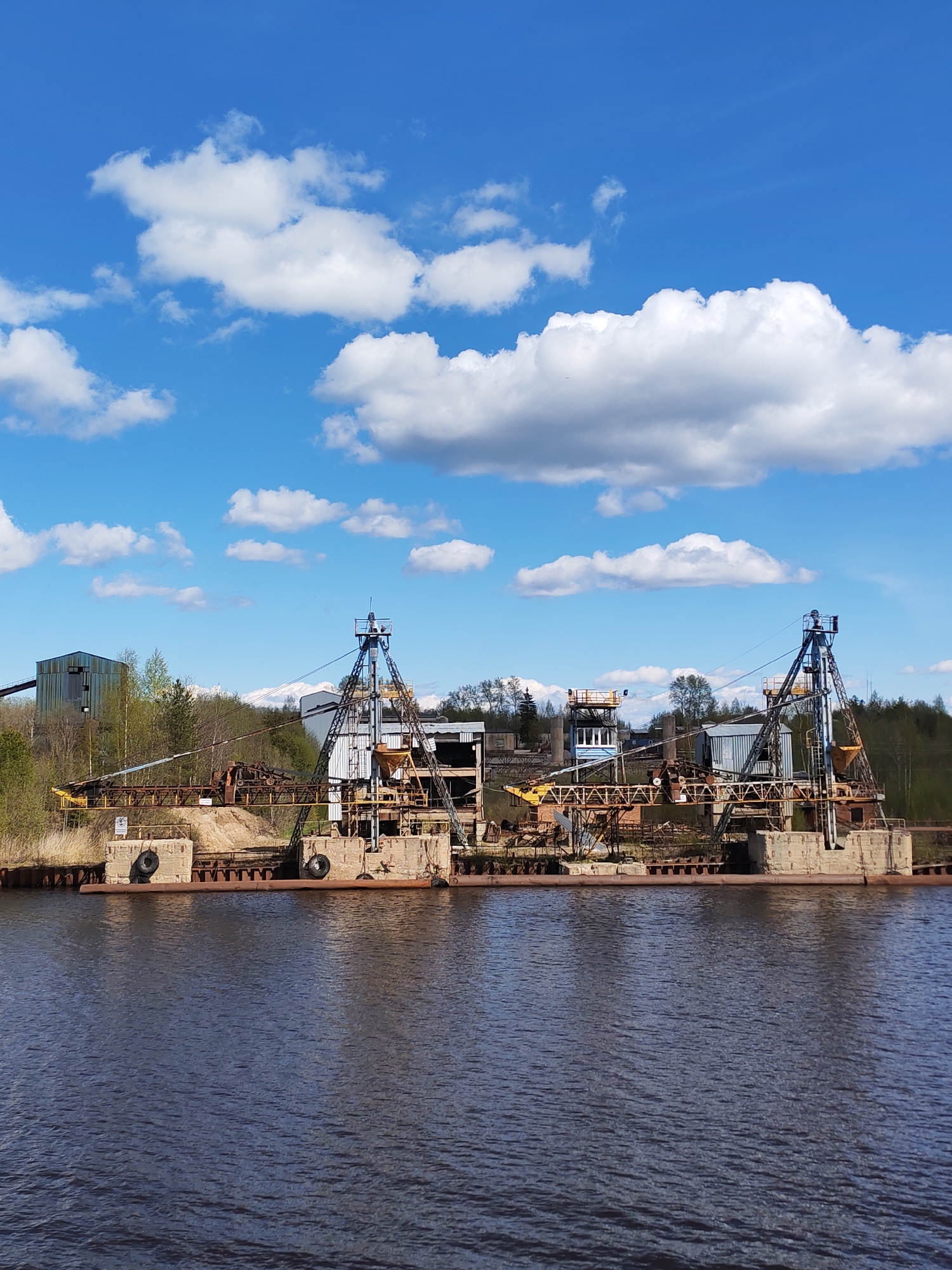 The image depicts an industrial waterfront scene in Ivanov Bor, Russia. Two large, decaying crane structures with rusted metal frames and concrete bases are situated on what appears to be a dilapidated dock or platform extending into a body of dark, rippling water. Old tires are fastened to the dock's edge. Behind the cranes, amidst vegetation and trees, are other industrial buildings, including a structure with a conveyor belt. The sky is bright blue with puffy white clouds. The overall impression is one of disuse, obsolescence, and industrial decline.