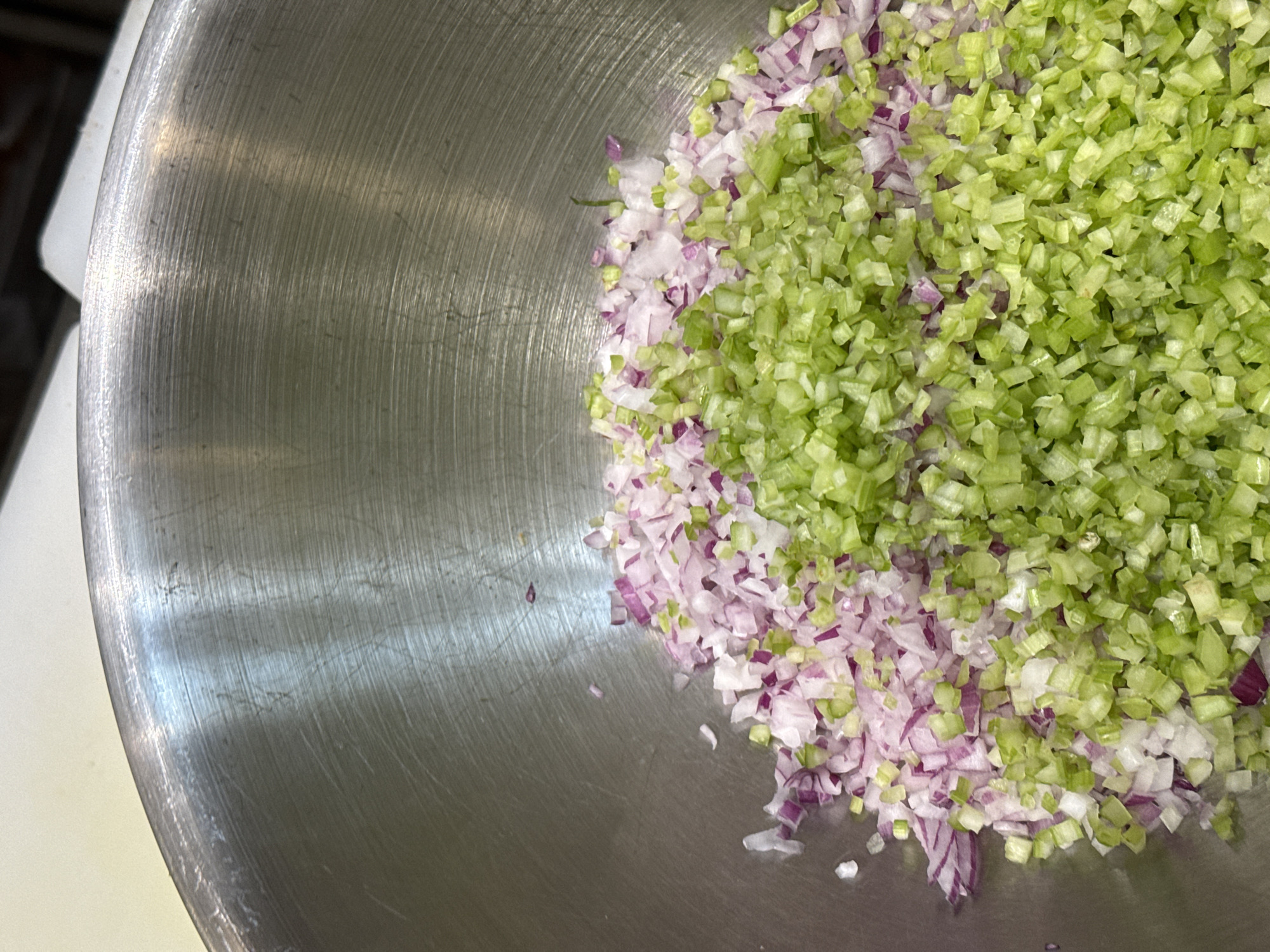 The image shows a close-up, overhead view of a metal bowl filled with finely chopped red onion and celery. The vegetables are arranged in distinct piles, with the red onion on the left and the celery on the right, both filling the bottom half of the bowl. The bowl itself appears to be made of stainless steel and shows signs of use, with visible scratches and a brushed finish. The background is out of focus and appears to be a light-colored countertop. There is no discernible indication of the location within Tel-Aviv, Israel, or any people, actions, or interactions.
