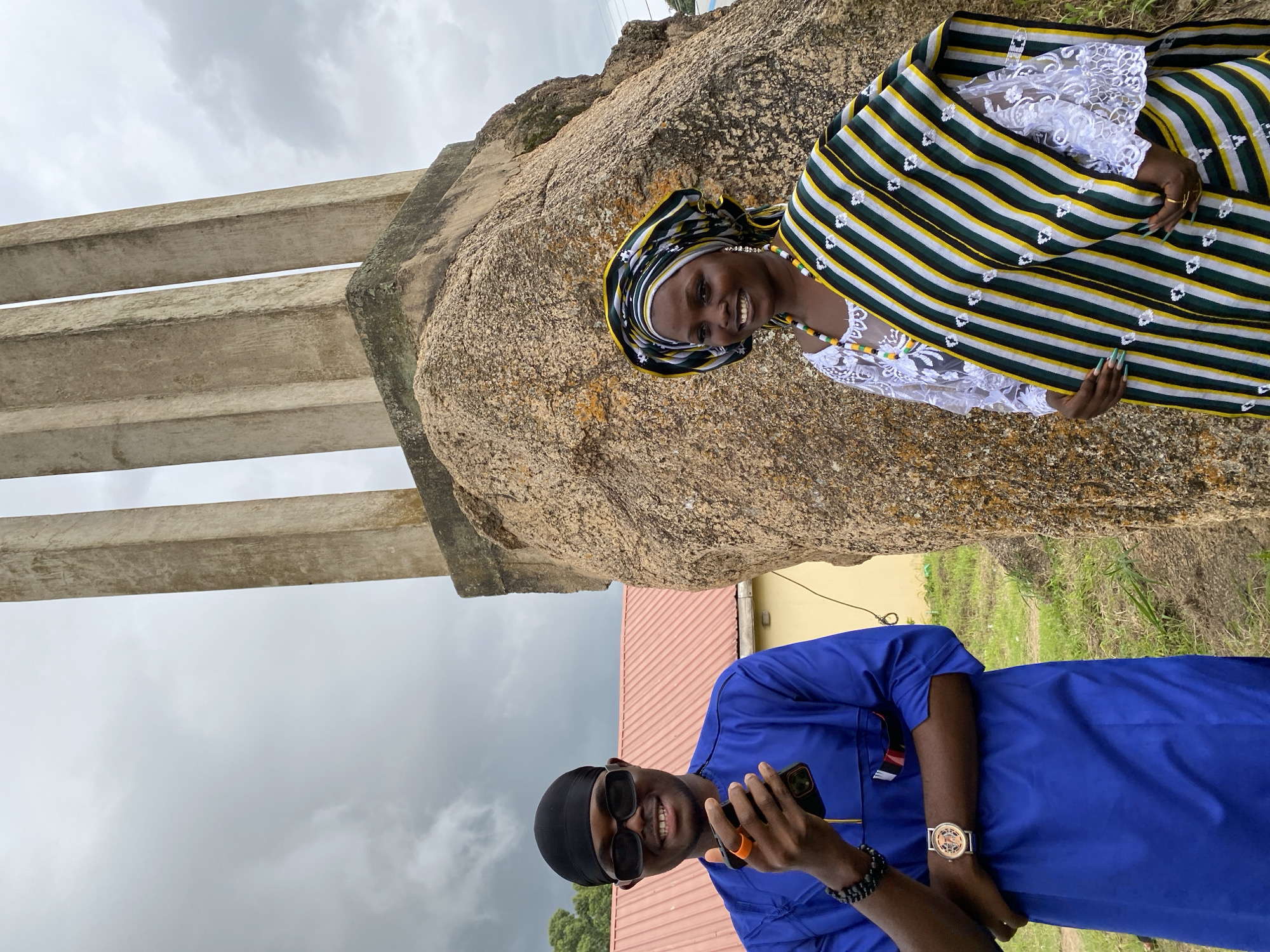 The media file features two individuals in traditional Nigerian attire, posing in an outdoor setting. The scene is dominated by a large rock formation, with several concrete pillars visible to the left, suggesting a historical or monumental site.
The woman, positioned slightly behind and to the right, wears a vibrant striped headscarf and a white lace top. She is adorned with a colorful beaded necklace. Her expression is a joyful smile, and she is looking towards the camera.
The man, in the foreground, is dressed in a royal blue garment. He is wearing dark sunglasses and a black head wrap. He is holding a smartphone in his right hand and has a wrist watch and bracelet on his left arm. He is also smiling and looking at the camera, with a relaxed and happy demeanor.
The background includes grassy ground and a building with a red roof. The sky is overcast and appears to be a cloudy day. The lighting suggests it is daytime, possibly late morning or afternoon. The overall atmosphere is positive and celebratory, with the subjects appearing to enjoy their time together at this location in Wang, Nigeria. There is no visible text in the media file.