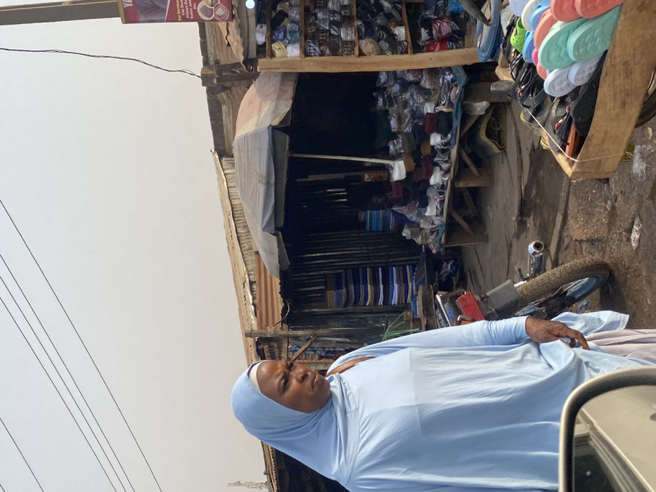 A woman in a light blue hijab and garment stands near a roadside shop. The shop displays stacks of various items, including footwear and fabric rolls, under a makeshift corrugated metal and tarp awning. A motorcycle is parked nearby. Power lines stretch across the sky. The scene appears to be in a bustling outdoor market area.