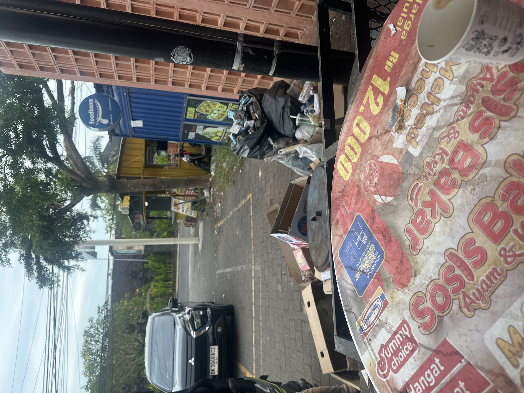 The image depicts a street scene in Celuk, Indonesia. A black car is parked along the side of the street with license plate "DK 1048 ACZ." In the background are businesses, including a Foodboxs restaurant, recognizable by its blue sign. Garbage is piled up on the side of the road, near some cardboard boxes. In the foreground is an outdoor table with a promotional sign for a food vendor, with logos for Indomaret and "Yummy choice," advertising "Sosis Bakar Ayam & Keju Melted Cheese" for Rp 12,000. A used paper cup sits on the table.