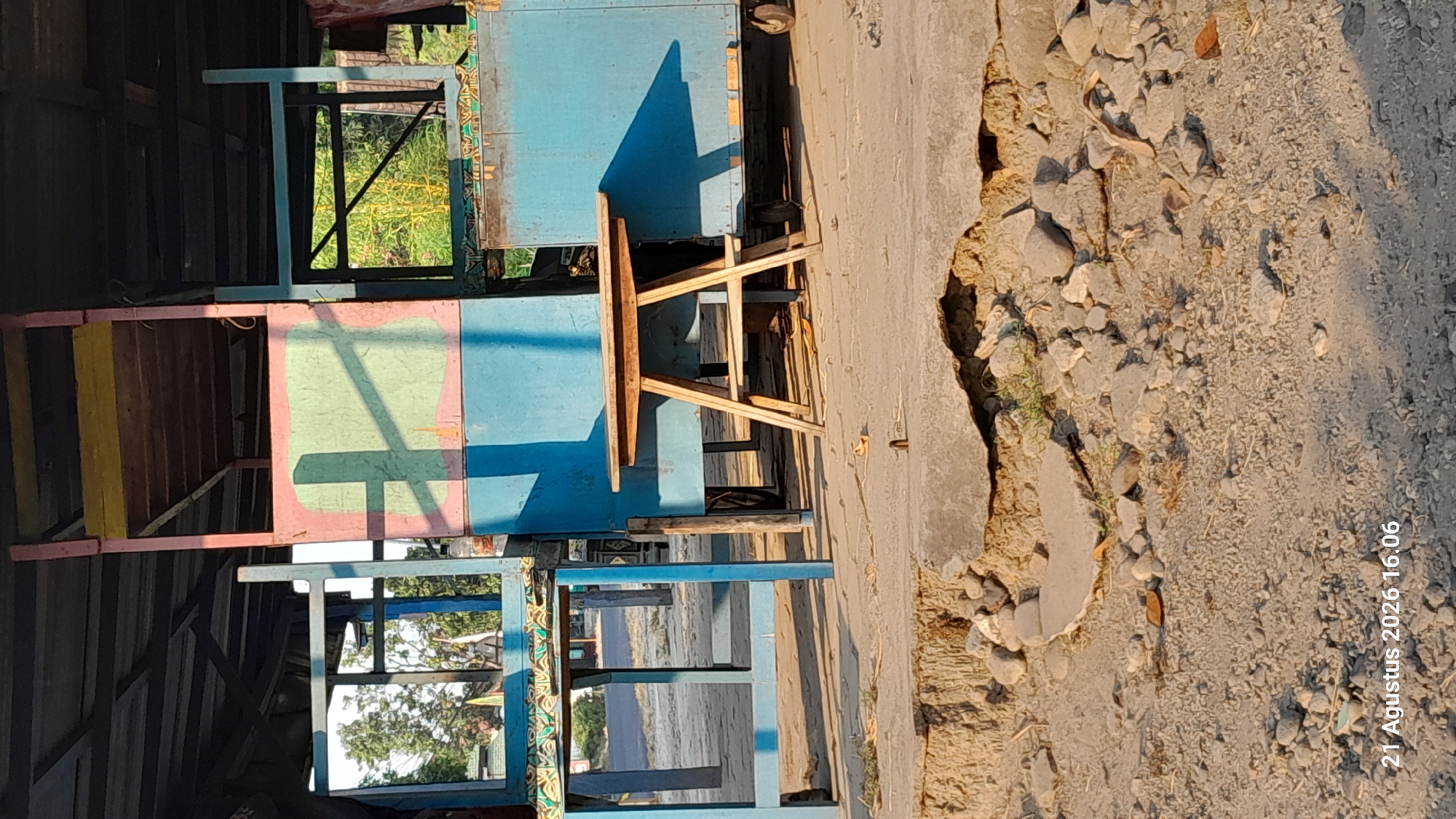 This image captures a scene in Takeran, Indonesia, focusing on an outdoor stall structure. The structure itself appears to be made of wood and painted in vibrant colors, including blue, pink, and yellow, with a green rectangular panel on one side. There is a small, light-colored wooden table with an A-frame base positioned in front of the stall. The ground in the foreground is uneven and consists of sand, pebbles, and broken concrete, suggesting a somewhat weathered or undeveloped area. In the background, through openings in the stall, trees and vegetation are visible, indicating an outdoor setting. The overall impression is that of a rustic, possibly abandoned or less-maintained stall in a tropical or subtropical environment.