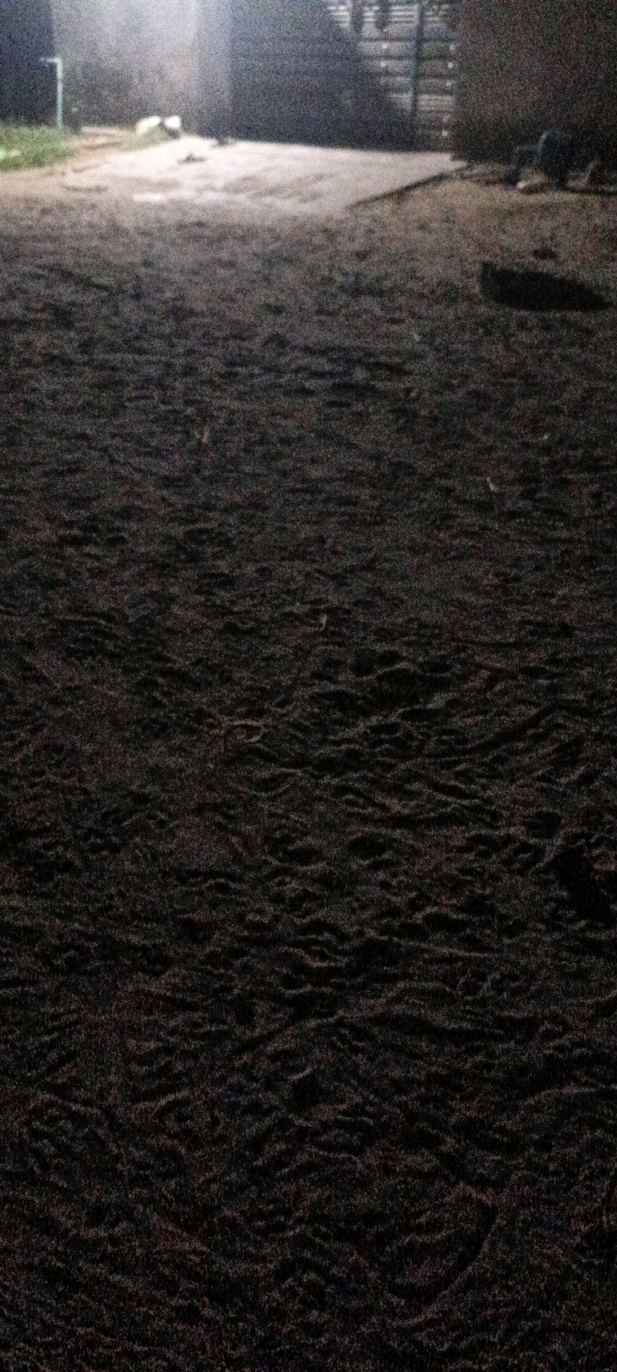 The image depicts a dark, rough, and uneven ground surface that appears to be dirt or coarse sand. The texture suggests it could be a pathway or an open area with windblown or trampled debris. In the background, a brighter, lighter area indicates a path or clearing illuminated by a light source. Structures are faintly visible in the far background, characterized by dark shapes and possibly some horizontal lines suggesting fencing or building materials. The overall lighting is dim, with the foreground being the darkest. The location is identified as CHAK NO.172 TDA, Pakistan.