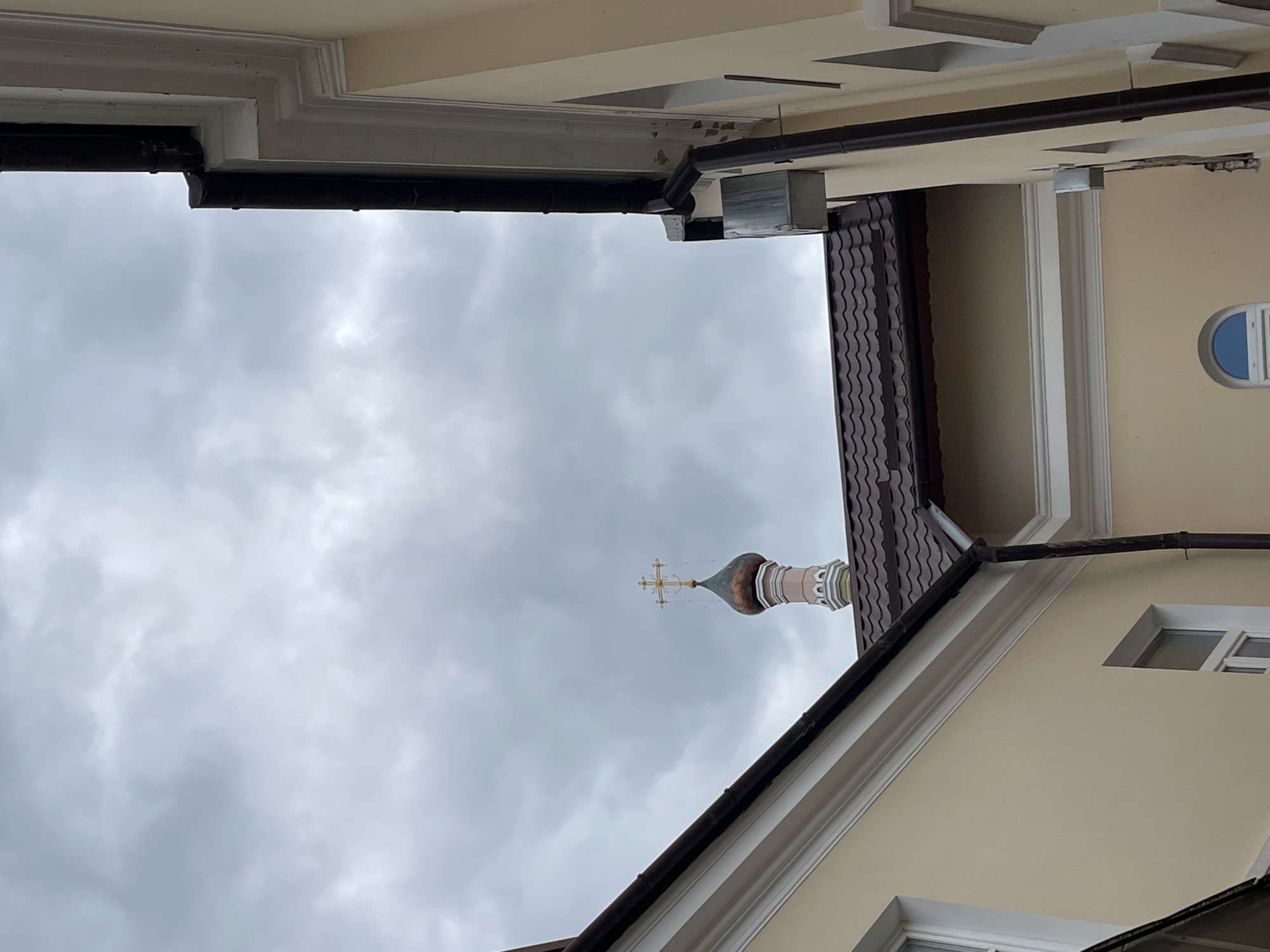 The image shows the exterior of a building with a church dome visible in the background. The architecture suggests a European style, possibly an Orthodox church given the dome's shape and the cross at its peak. The building in the foreground is painted in a light yellow hue and features white decorative trim around the windows and roofline. Dark brown gutters and downspouts are visible along the side of the building. The sky is overcast with gray clouds.
Based on the architectural style and the prominent church dome, this scene is likely located in Hrodna, Belarus, a city known for its historical churches and religious architecture. The specific location within Hrodna cannot be determined from the image alone.