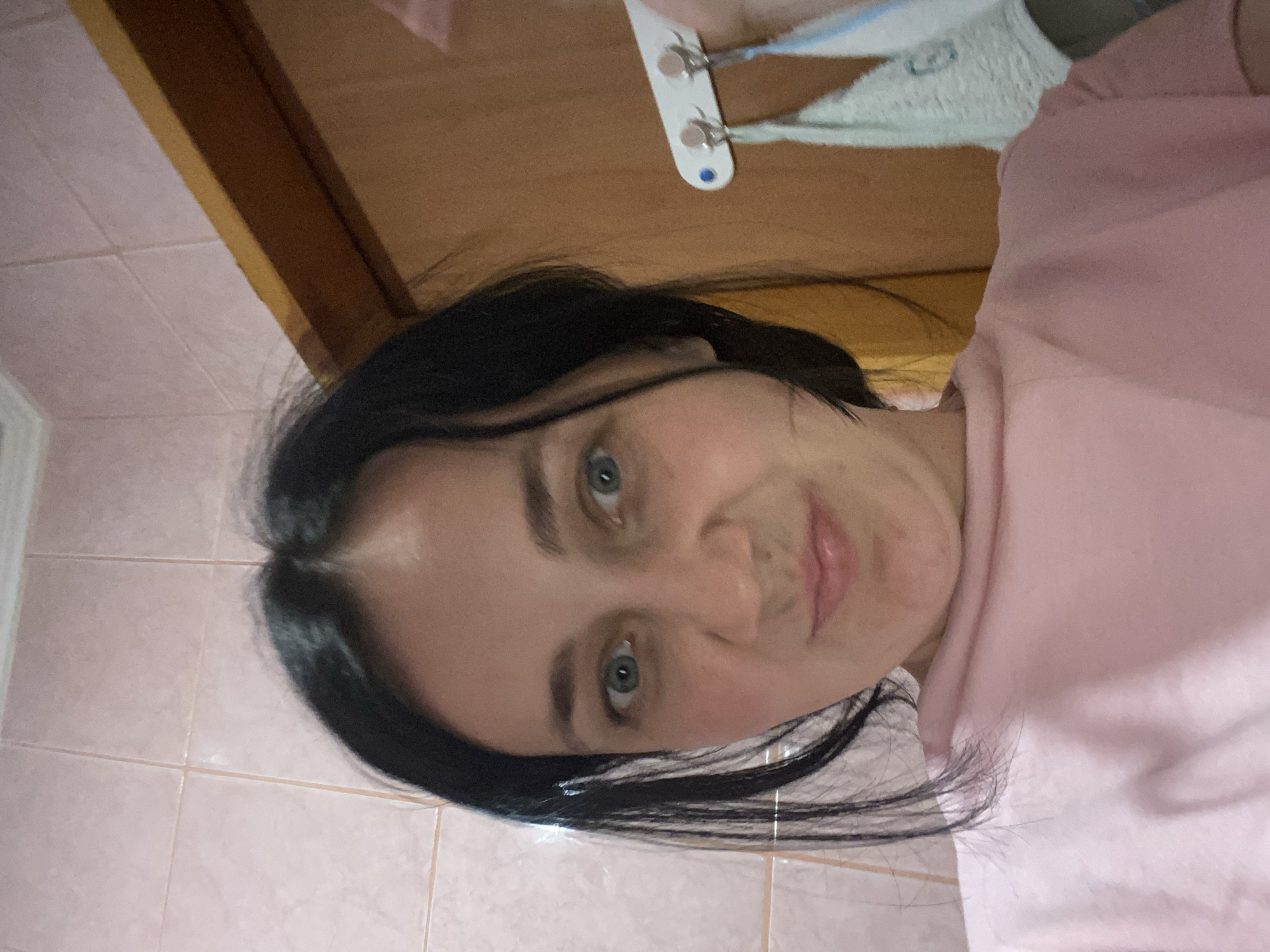 A young woman with dark hair and blue eyes is pictured indoors, likely in a bathroom, given the tiled walls. She is wearing a pink t-shirt. Her expression is a slight grimace, with her brow furrowed and her mouth turned down at the corners. To her right, a wooden doorframe and a towel rack with a light-colored towel are visible. The lighting is dim, and the overall atmosphere is casual.