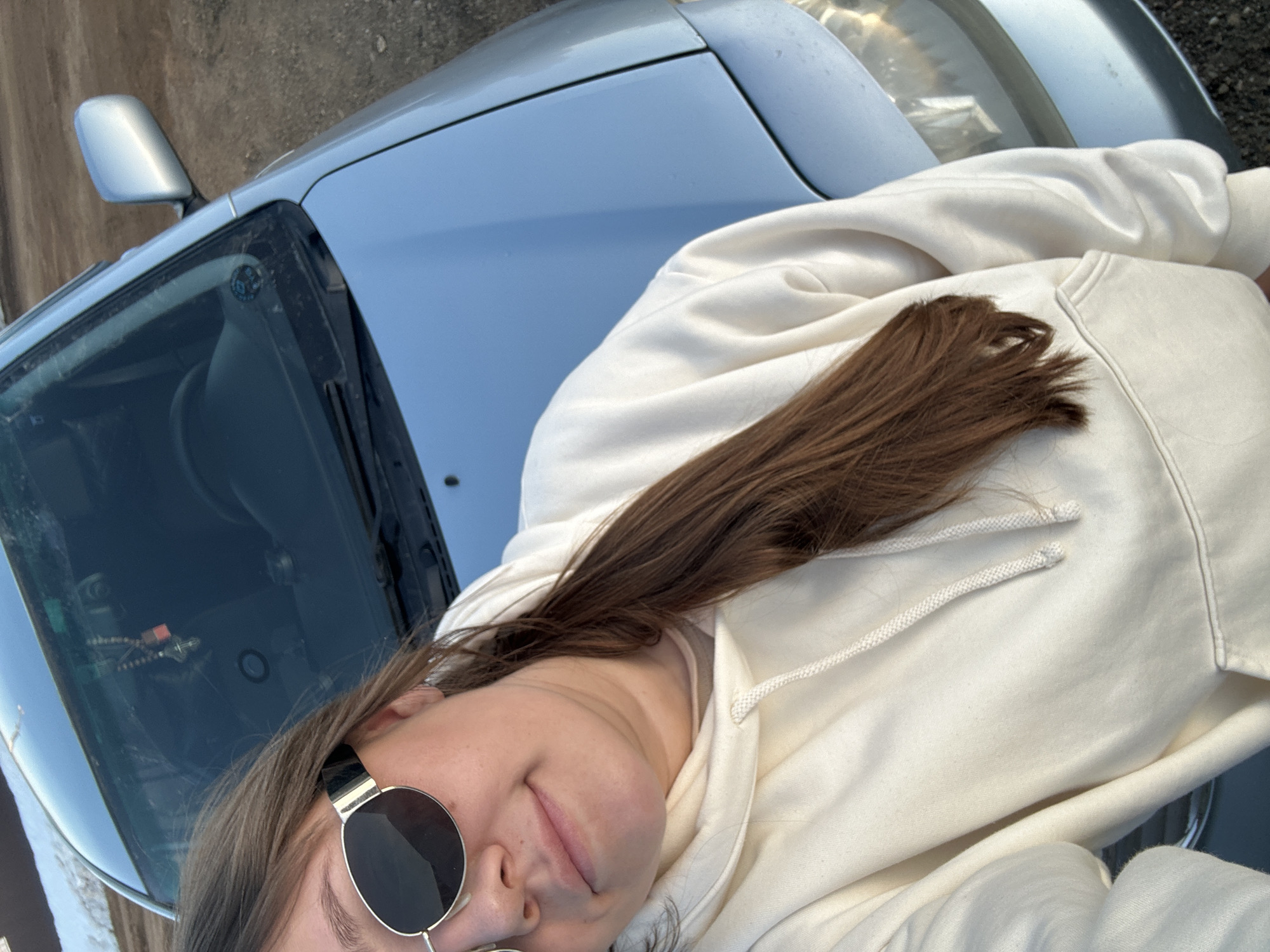 A young woman lies on the hood of a silver car, wearing round sunglasses and a cream-colored hoodie. Her brown hair rests on the hoodie. The car's windshield and side mirror are visible. The location appears to be outdoors. The city, Ольховка, Russia, is inferred from the image description request and is not visible in the image.