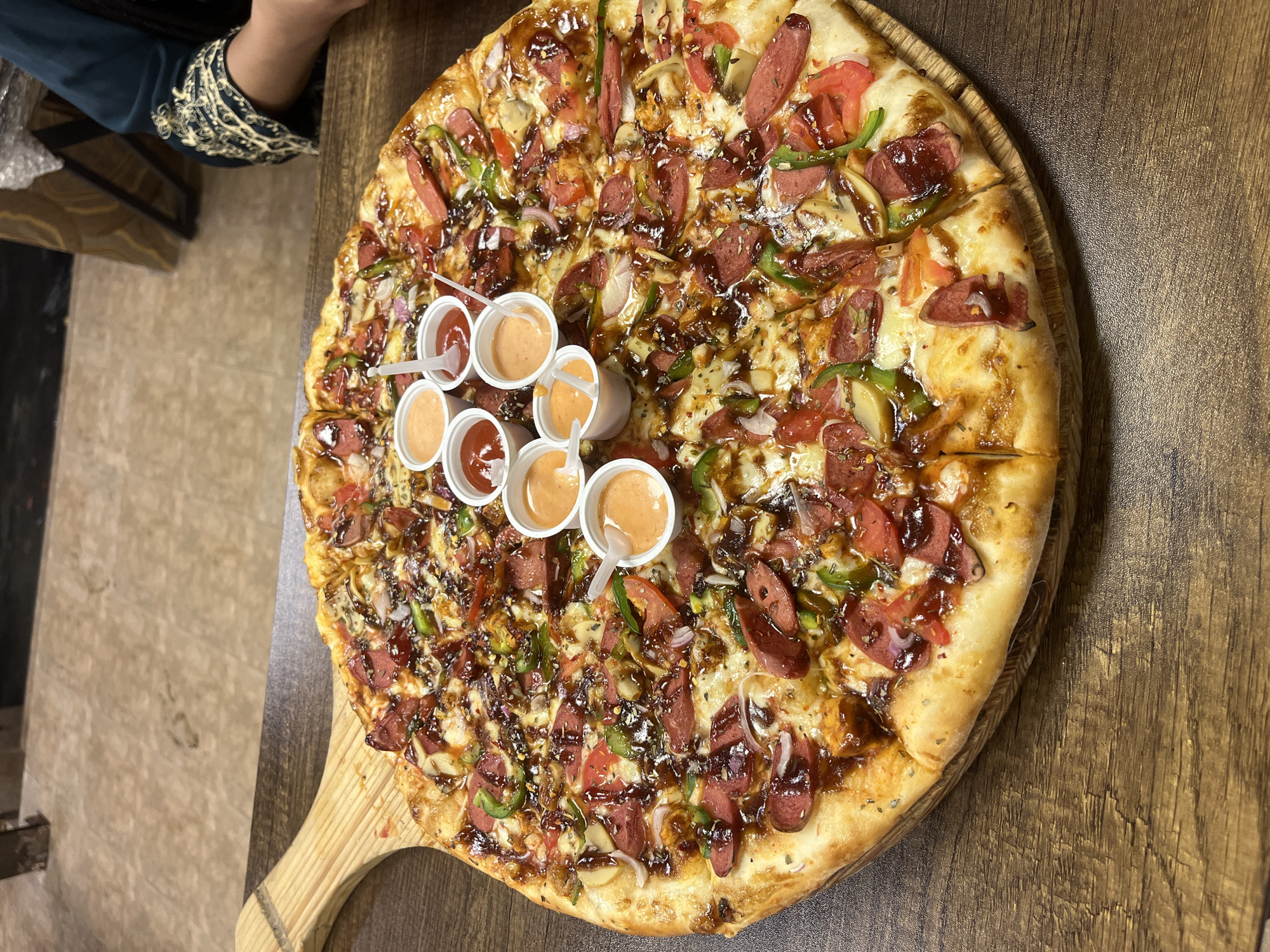 A pizza topped with sliced sausage, green bell peppers, sliced tomatoes, onions, mushrooms, and a dark sauce sits on a wooden paddle on a wooden table. Seven small plastic cups of sauce are arranged in the center of the pizza. A person's hand and decorated sleeve are visible in the upper left corner. The setting may be a restaurant in Dhaka, Bangladesh, based on the image's origin.