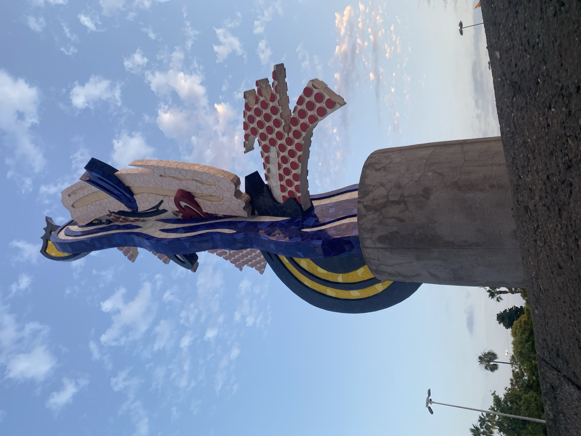 The image shows a low-angle view of the "Dona i Ocell" (Woman and Bird) sculpture by Joan Miró, located in Barcelona, Spain. The abstract sculpture is brightly colored, featuring vibrant blues, yellows, reds, and whites, with a mosaic tile finish. It stands on a cylindrical concrete base. The sculpture is situated in a park-like setting with a clear blue sky dotted with white clouds in the background. To the left and in the foreground, there are palm trees and other greenery, along with streetlights. The foreground also features a dark, textured surface, possibly a wall or embankment. The lighting suggests it is either early morning or late afternoon.