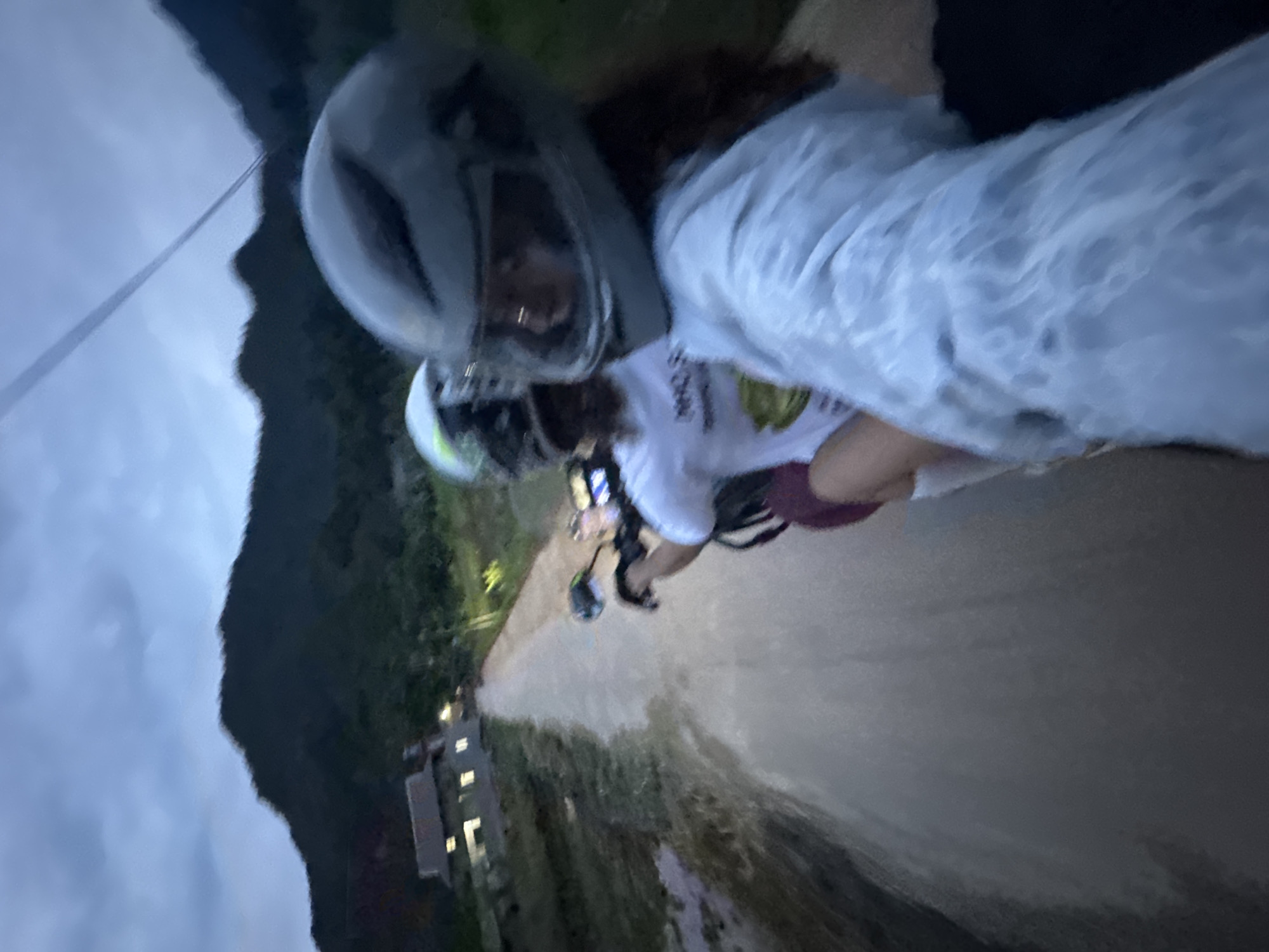 The image captures a low-angle, slightly blurred view from the perspective of someone riding on the back of a motorcycle, likely at dusk or early evening. Two individuals are visible, both wearing helmets. The person in the foreground, with a gray helmet, is wearing a light blue, possibly tie-dyed, t-shirt and has dark hair. Their right arm is visible, holding onto something, and their face is turned slightly away from the camera. Behind them, another person in a white t-shirt with a graphic on it and a lighter-colored helmet is seated in front.
They are traveling on a dirt or unpaved road that stretches into the distance. On the left side of the road, there's a traditional Vietnamese house with illuminated windows. Lush vegetation and dark, undulating hills or mountains form the backdrop. The sky is overcast and a deep blue-gray, suggesting twilight or cloudy conditions. The overall impression is of a rural setting in Mai Chau, Vietnam, with the journey taking place along a quiet, scenic route.