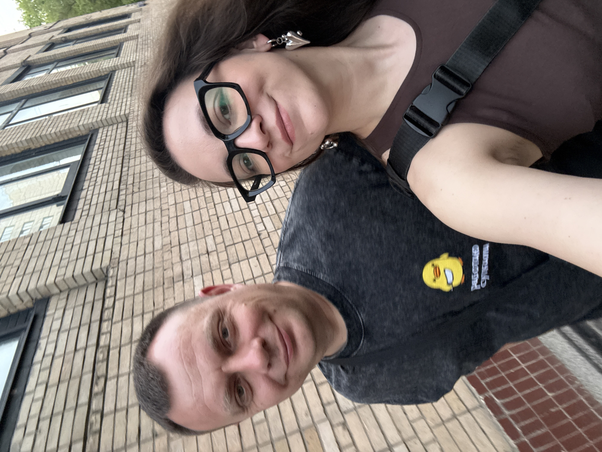 The image shows a selfie of two people, a man and a woman, seemingly standing on a street. The woman is at the top of the frame wearing glasses, a brown top, and a silver earring. The man is at the bottom, wearing a dark gray t-shirt with a small yellow duck cartoon on it. The background shows a building with multiple windows and a brick pattern, indicating an urban setting. The ground appears to be paved. Given the duck cartoon on the man's shirt (inscribed with Russian characters), and other context clues, it is plausible that the location is Moscow, Russia.