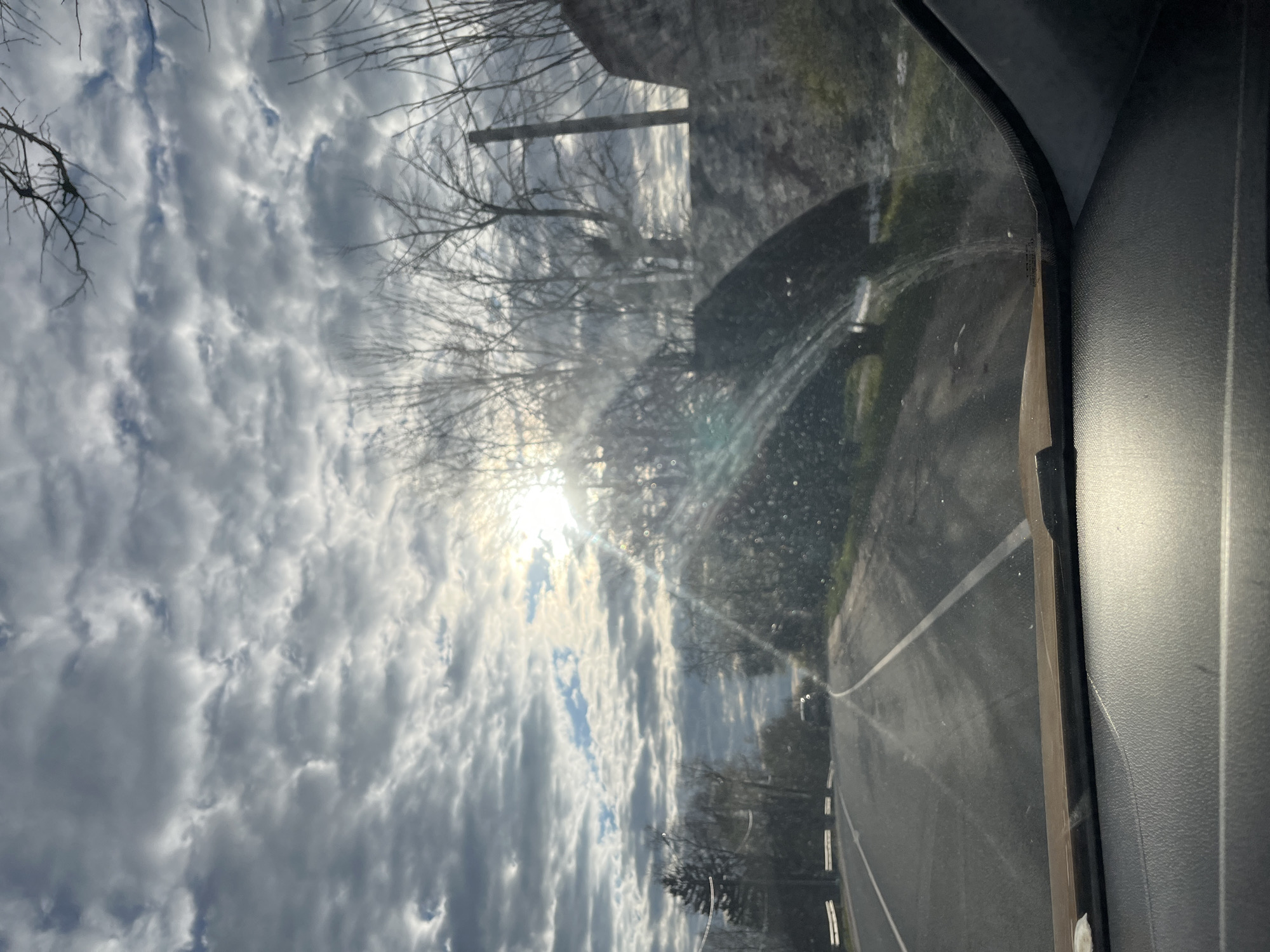 The image, captured in Maksimkovo, Russia, is taken from inside a vehicle looking out through the windshield. The original photograph is rotated 90 degrees clockwise. When viewed with the sky at the top and the road at the bottom, the scene unfolds during daytime, likely morning or late afternoon, under partly cloudy conditions.
The upper portion of the frame is dominated by a textured, partly cloudy sky, featuring dense, grey and white stratocumulus or altocumulus clouds interspersed with brighter patches of clear sky. A prominent sunburst emanates from the upper-left, partially obscured by clouds, creating significant glare and lens flares across the upper-middle section of the image, indicating the sun's intensity.
Silhouetted against the bright sky are numerous bare branches of deciduous trees, suggesting a winter or early spring setting. In the mid-ground, along the sides of the road, more densely vegetated areas and possibly distant structures are visible, though somewhat indistinct due to the lighting and distance.
A paved road occupies the lower-middle portion of the image, stretching into the distance. It is marked with white lane lines, possibly a double solid line or a single line along its right edge, and its surface appears somewhat reflective.
On the far right, elements of the car's interior are visible, including a dark A-pillar and a portion of the textured dashboard or door panel, confirming the perspective from within a car. The windshield through which the photo was taken shows numerous small specks and streaks, likely dirt or water droplets, which contribute to the diffused light and scattered appearance of the glare.
The image captures a moment of travel, as if driving, with no discernible people or specific event taking place. There is no visible text in the image.