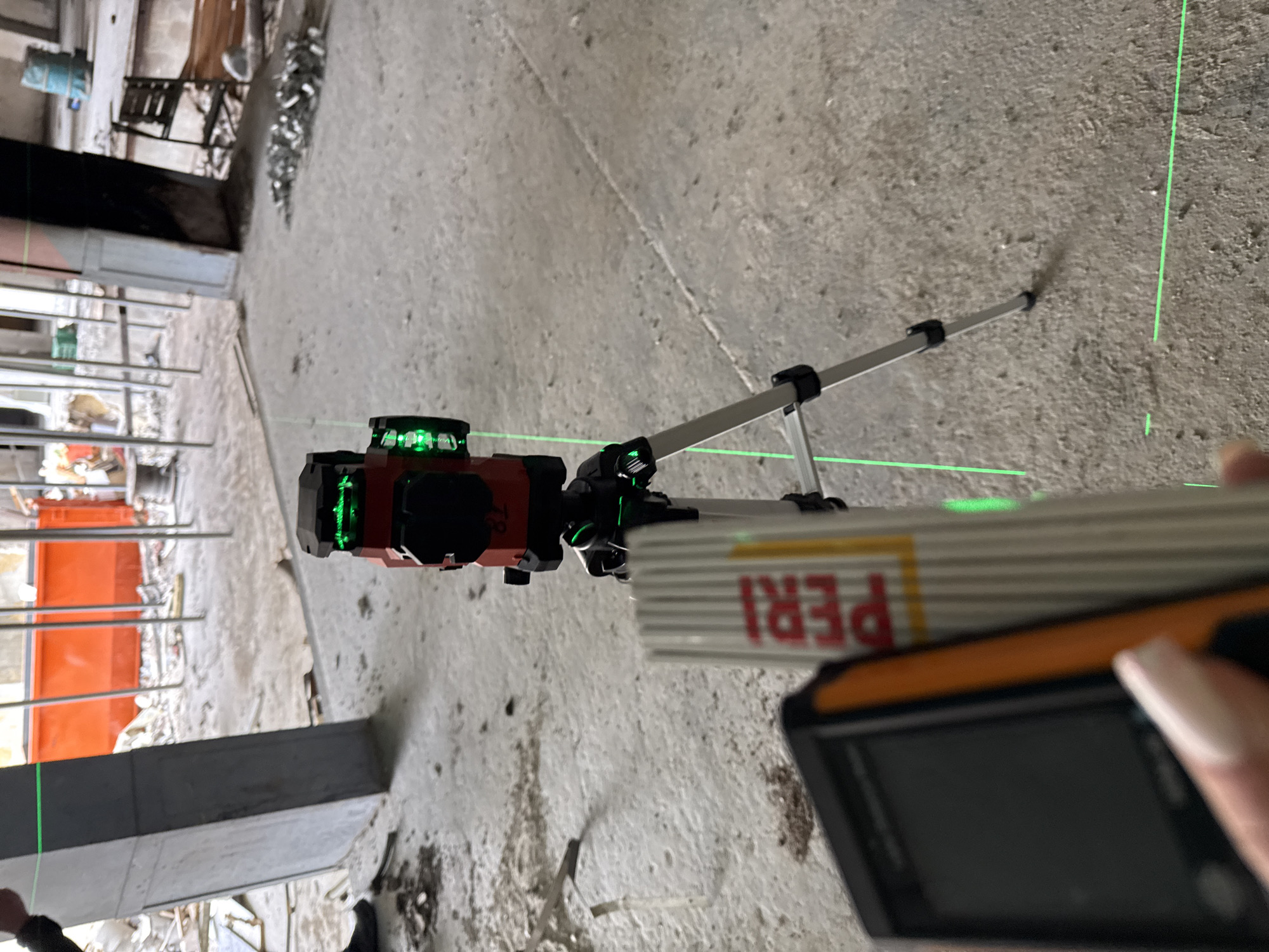 The image shows a construction site, potentially in Varna, Bulgaria, given the "PERI" brand on the measuring device. A laser level on a tripod projects green laser lines across the concrete floor and onto nearby structures. A person is holding a handheld laser distance meter, seemingly taking measurements. The environment includes exposed rebar, scaffolding, and other construction debris, suggesting an active or recently active building project.