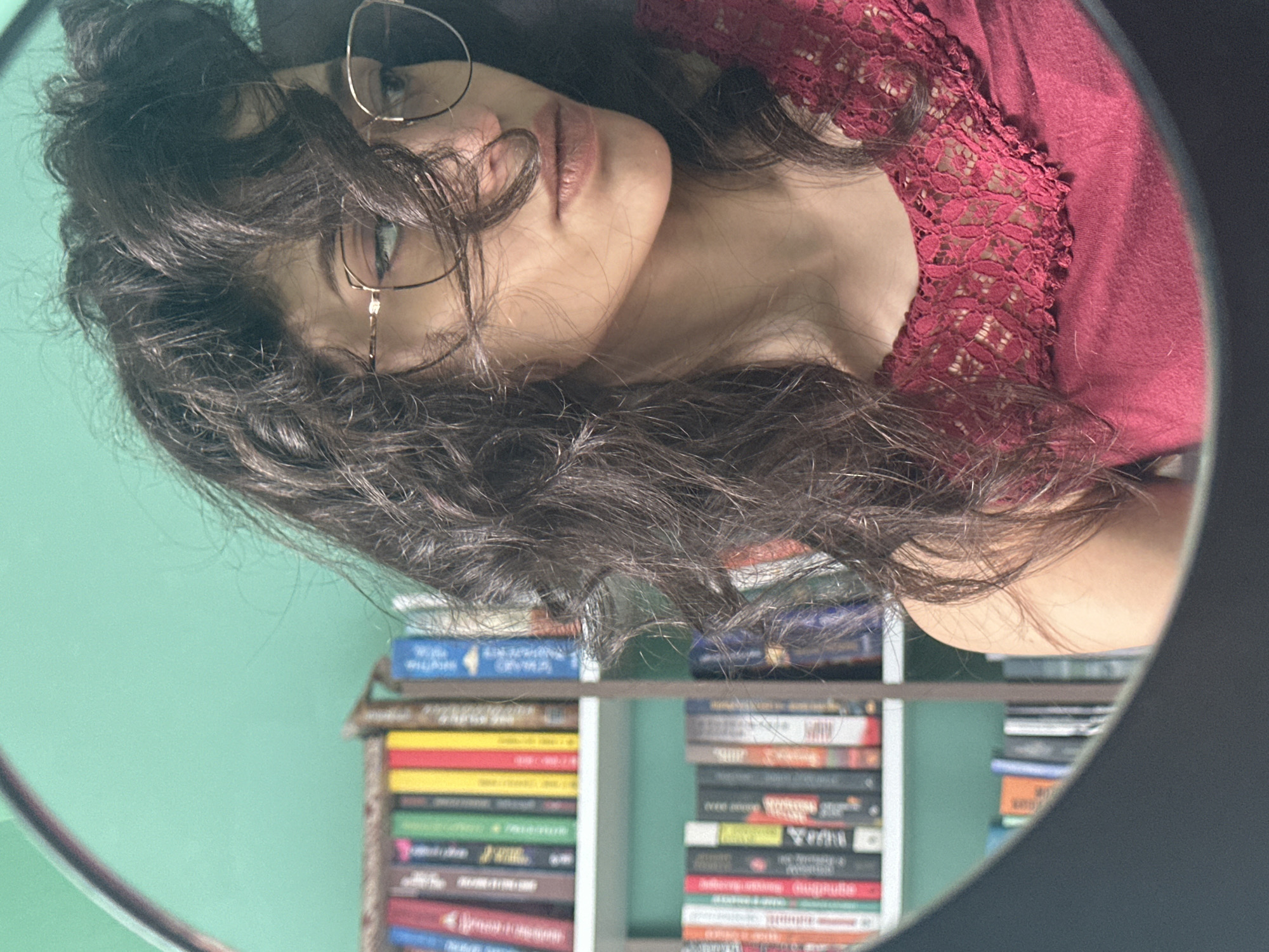 The image shows a reflection of a young woman wearing glasses and a maroon top with lace detailing. She has dark, curly hair and is looking away from the camera. The background consists of bookshelves filled with books, set against a mint green wall. The scene appears to be an indoor setting, possibly a home library or study. There is no information in the image to suggest a location in Sofia, Bulgaria.