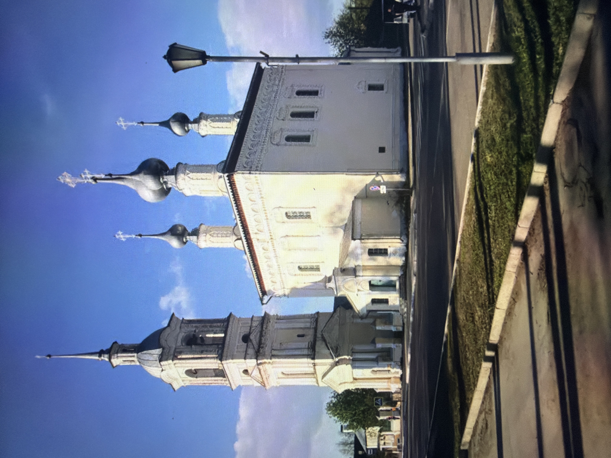 The image captures a clear, sunny day in Kotelniki, Russia, featuring the Church of the Archangel Michael. The architecture of the church is predominantly white with distinctive onion domes topped with crosses. A tall, slender bell tower stands to the left of the main building. The scene is viewed from across a paved area, likely a street or plaza, with a strip of grass and a curb separating it from the church. A lamppost stands on the right side of the frame. The sky is a vibrant blue with a few scattered white clouds. In the background, there are hints of other buildings and trees, as well as a few people walking and a vehicle. The lighting suggests it is daytime, possibly late afternoon, given the long shadows cast on the ground.